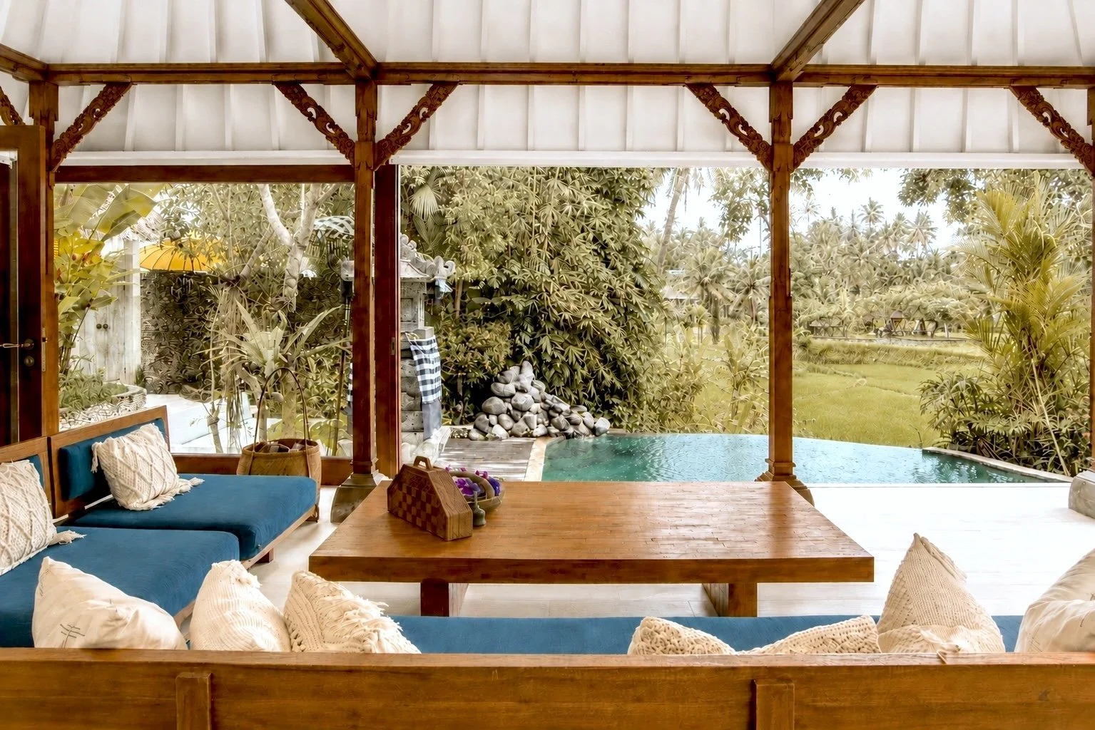 Indoor seating area with blue cushions and white pillows, wooden table, overlooking a lush outdoor garden with trees, plants, rocks, a small pool, and a waterfall feature.