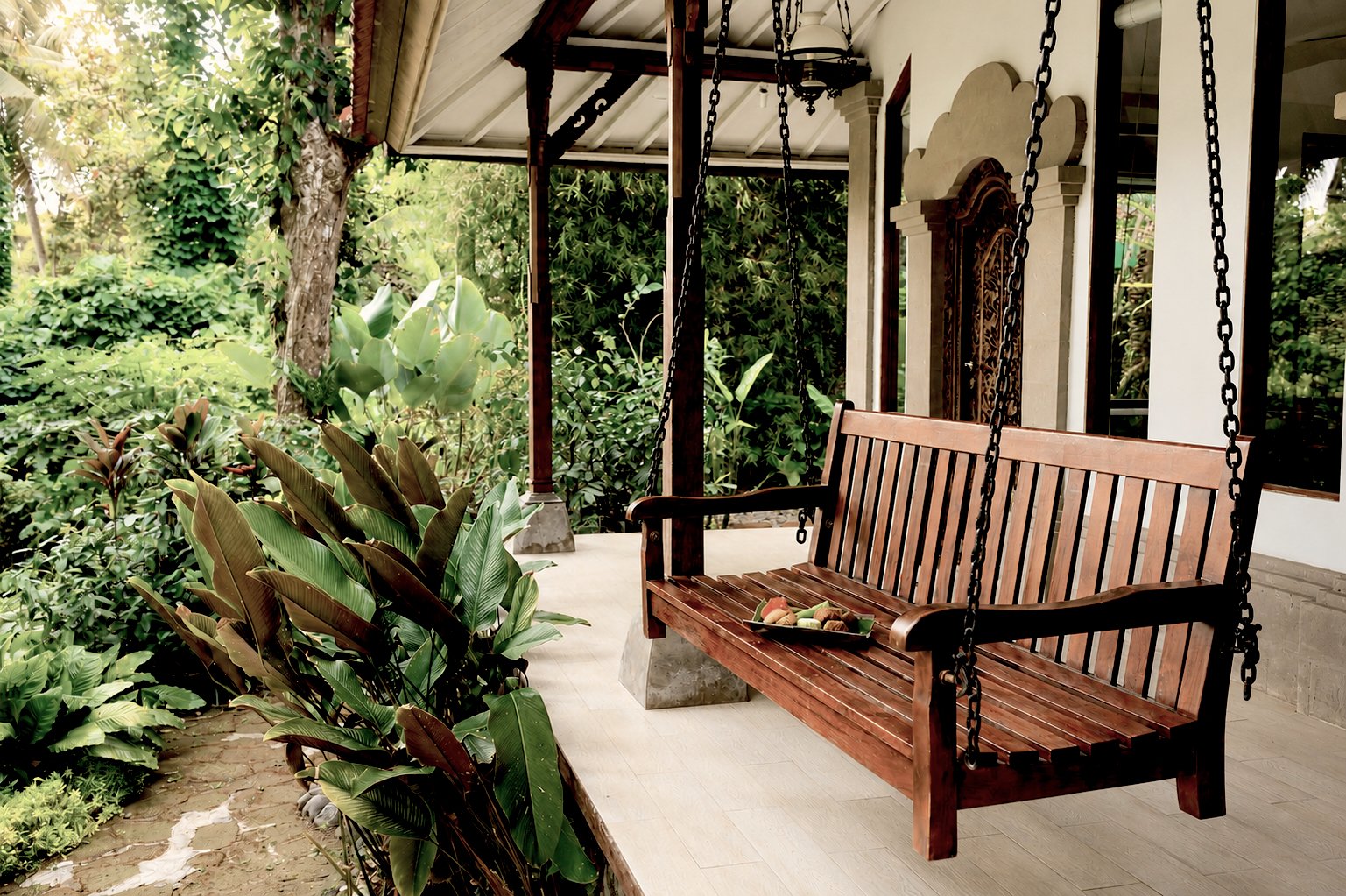 A porch with a wooden swing hanging from chains, surrounded by green plants and trees, with a tray of fruit on the swing.