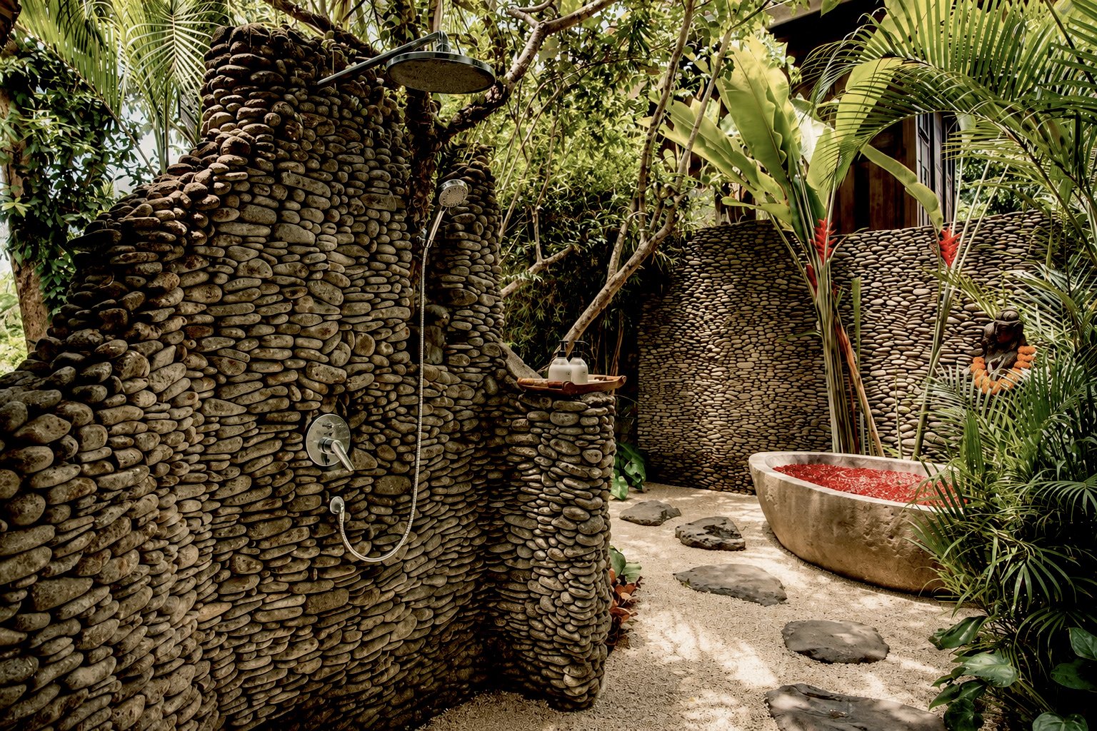 Outdoor spa area with stone walls, a large stone bathtub filled with red water, and lush tropical plants surrounding the space.