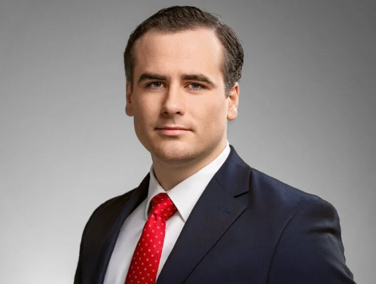A professional portrait of a man in a dark suit, white shirt, and red tie with a subtle pattern, posing against a gray background.