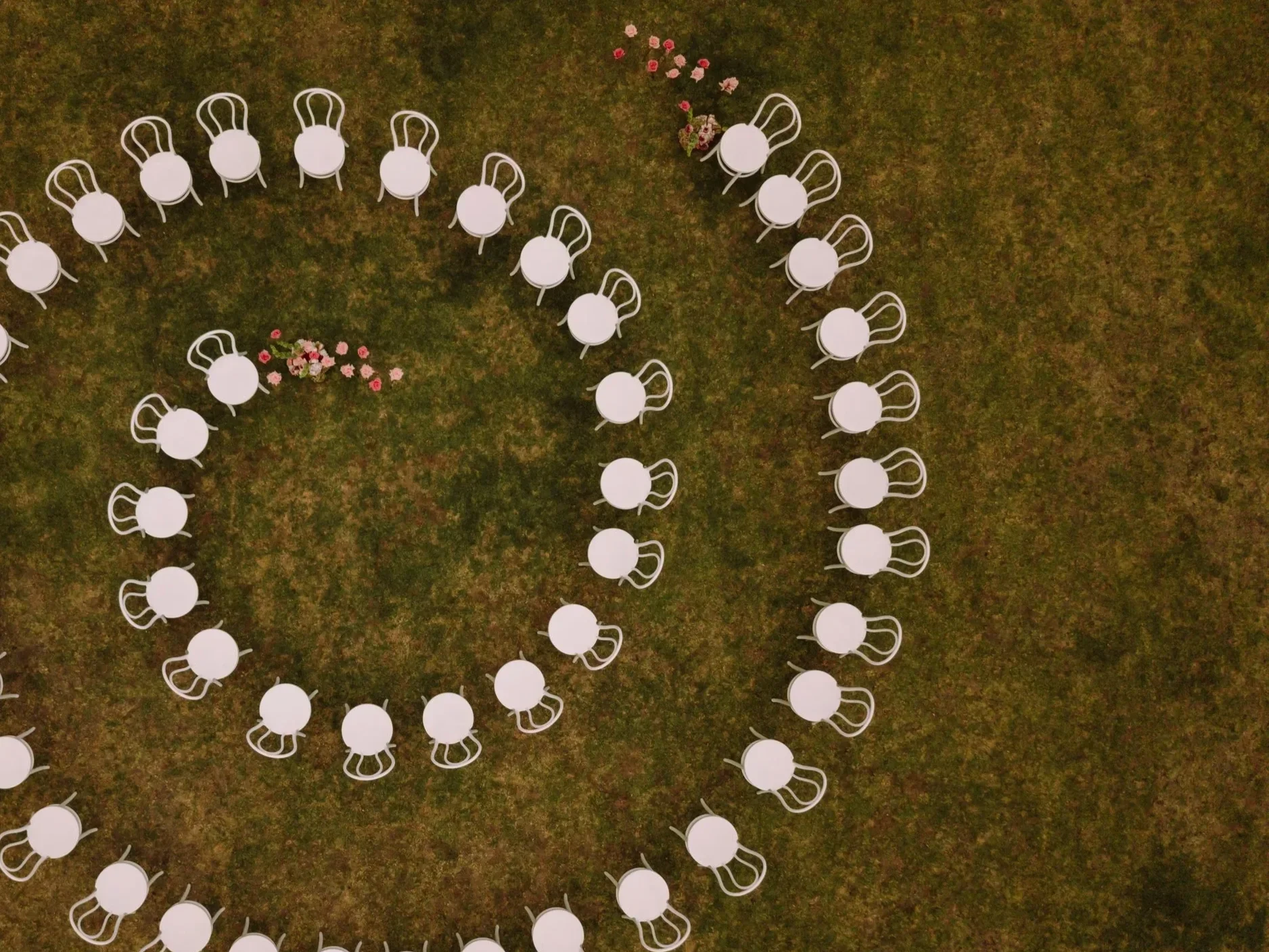 Overhead view of white round tables and chairs arranged in concentric circles on grass, with a small cluster of pink and white flowers.