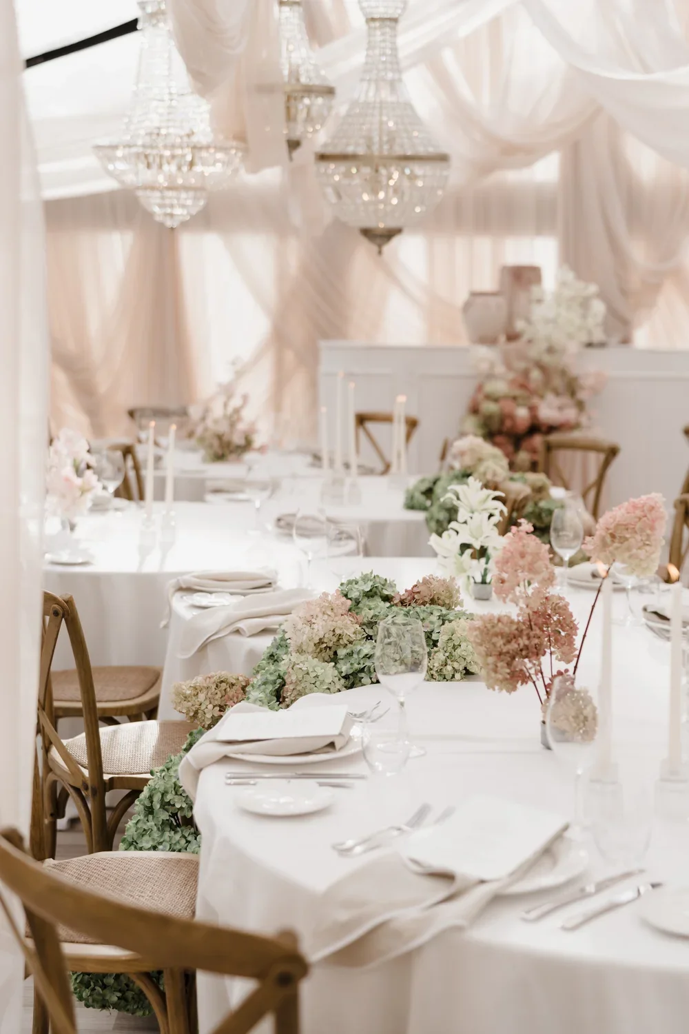 Elegant marquee wedding reception with draped white fabric, crystal chandeliers, and tables decorated with floral centerpieces and place settings designed and styled by Alex Morabito and Ruffles and Bells.