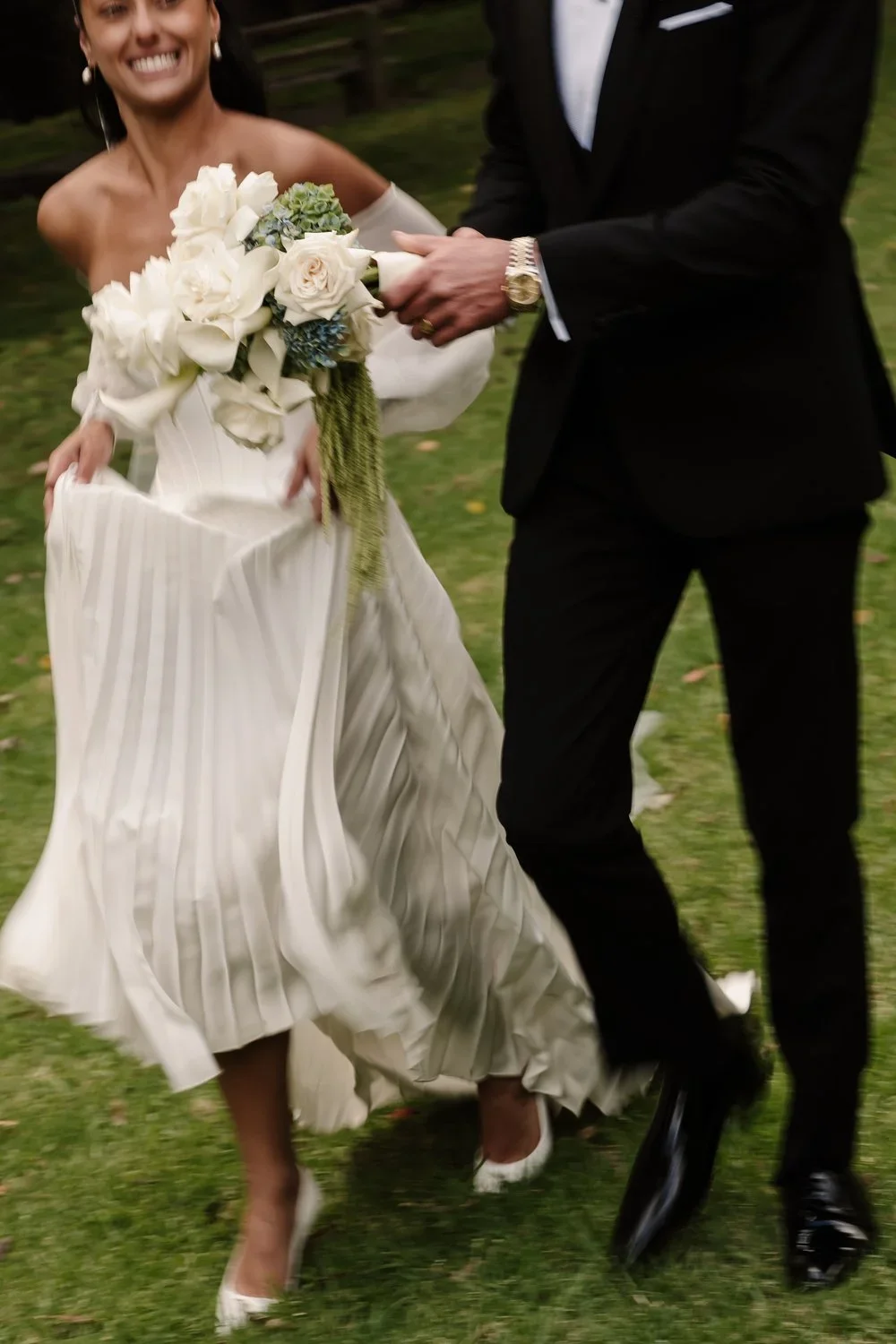Luxury Bride in a white wedding gown holding a bouquet, smiling and walking on grass with groom in a black tuxedo, in an outdoor wedding setting in Red Hill on the Mornington Peninsula.