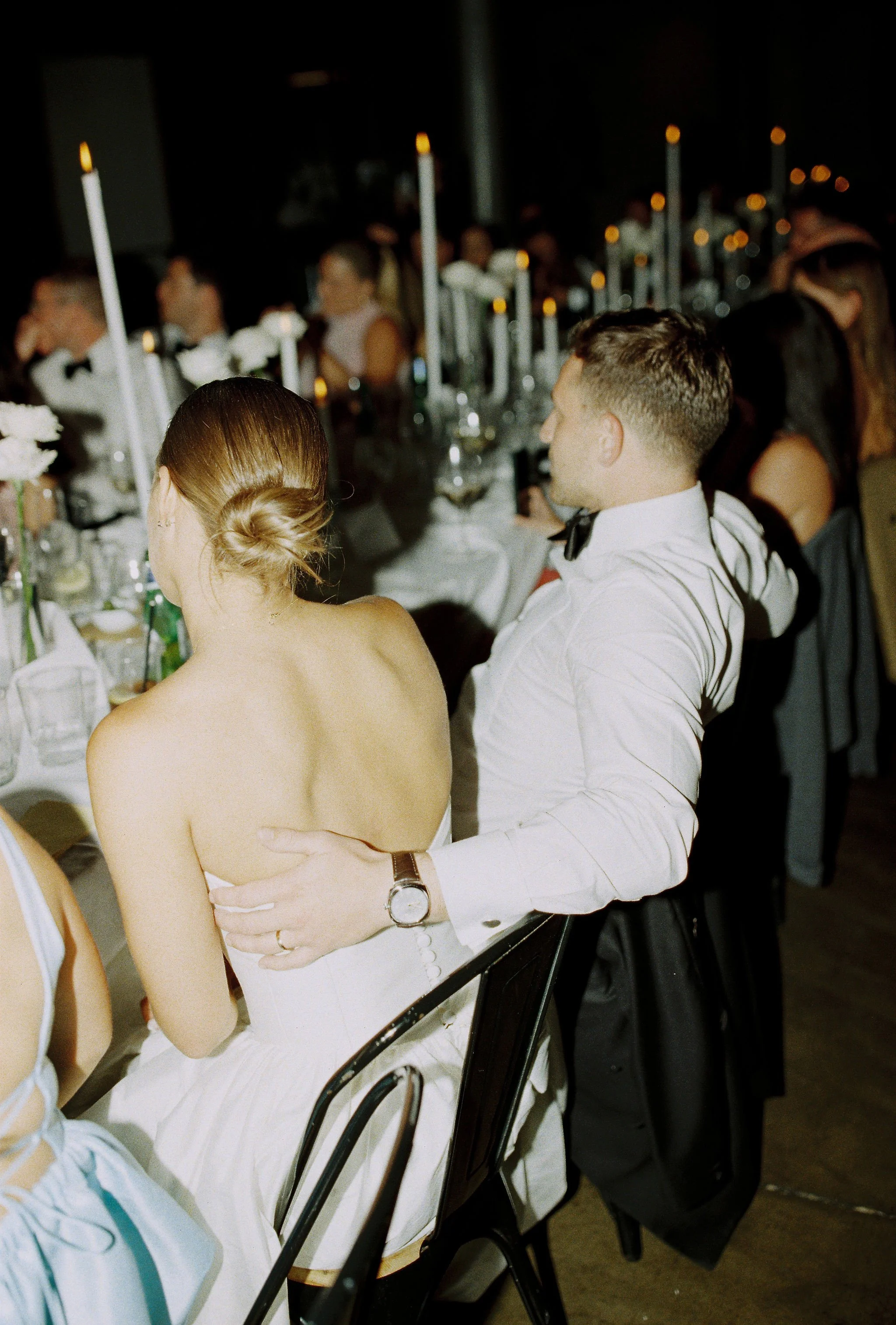 A couple at a formal event, the man in a white dress shirt with a bowtie and the woman with her back turned, displaying an off-the-shoulder dress and her hair in a low bun, sitting at a decorated banquet table with candles and flowers.