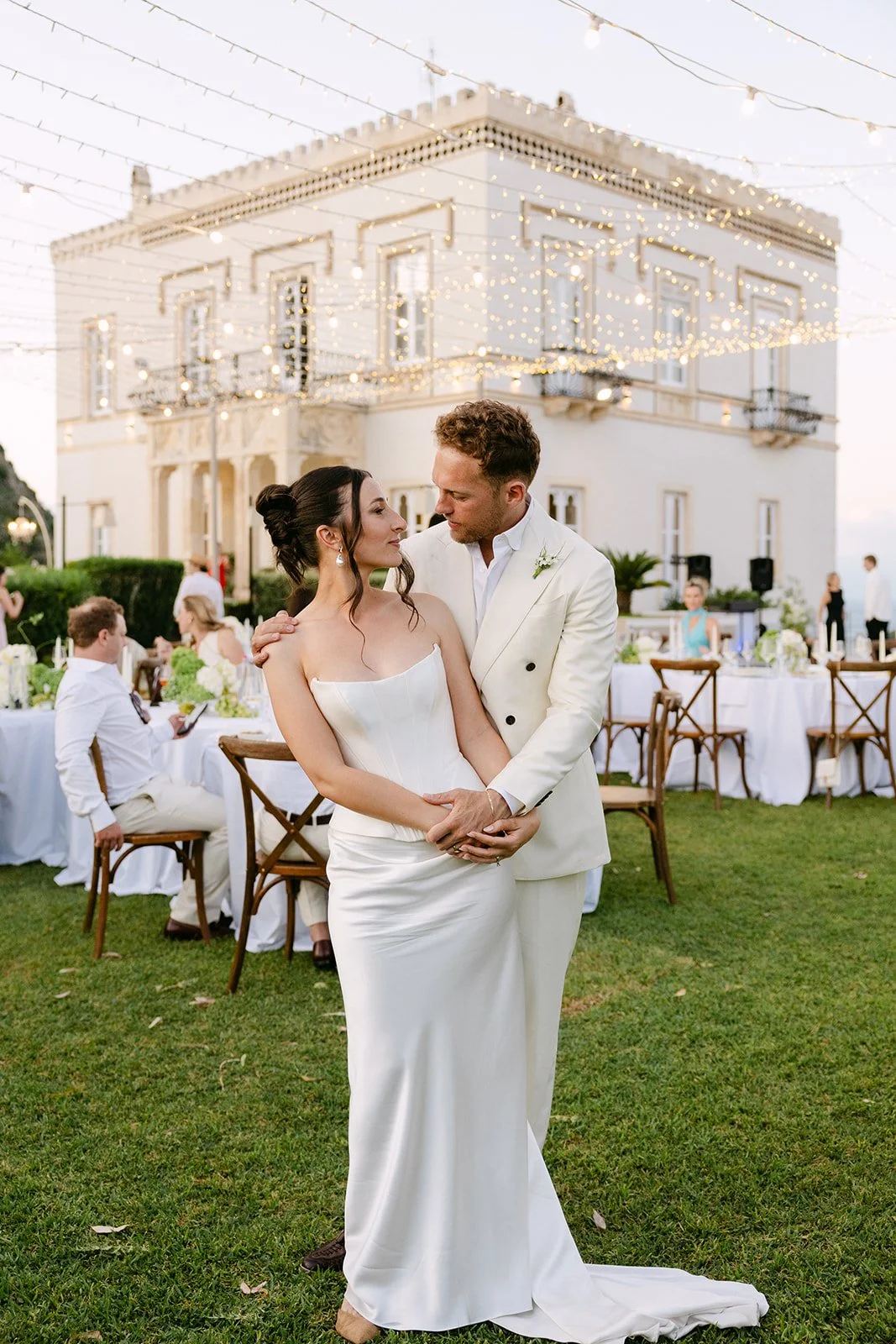 A bride and groom share an intimate moment at their outdoor Italian destination wedding at Villa Mon Repos in Taormina, under string lights on a lawn, with guests seated at tables styled by Alex Morabito.