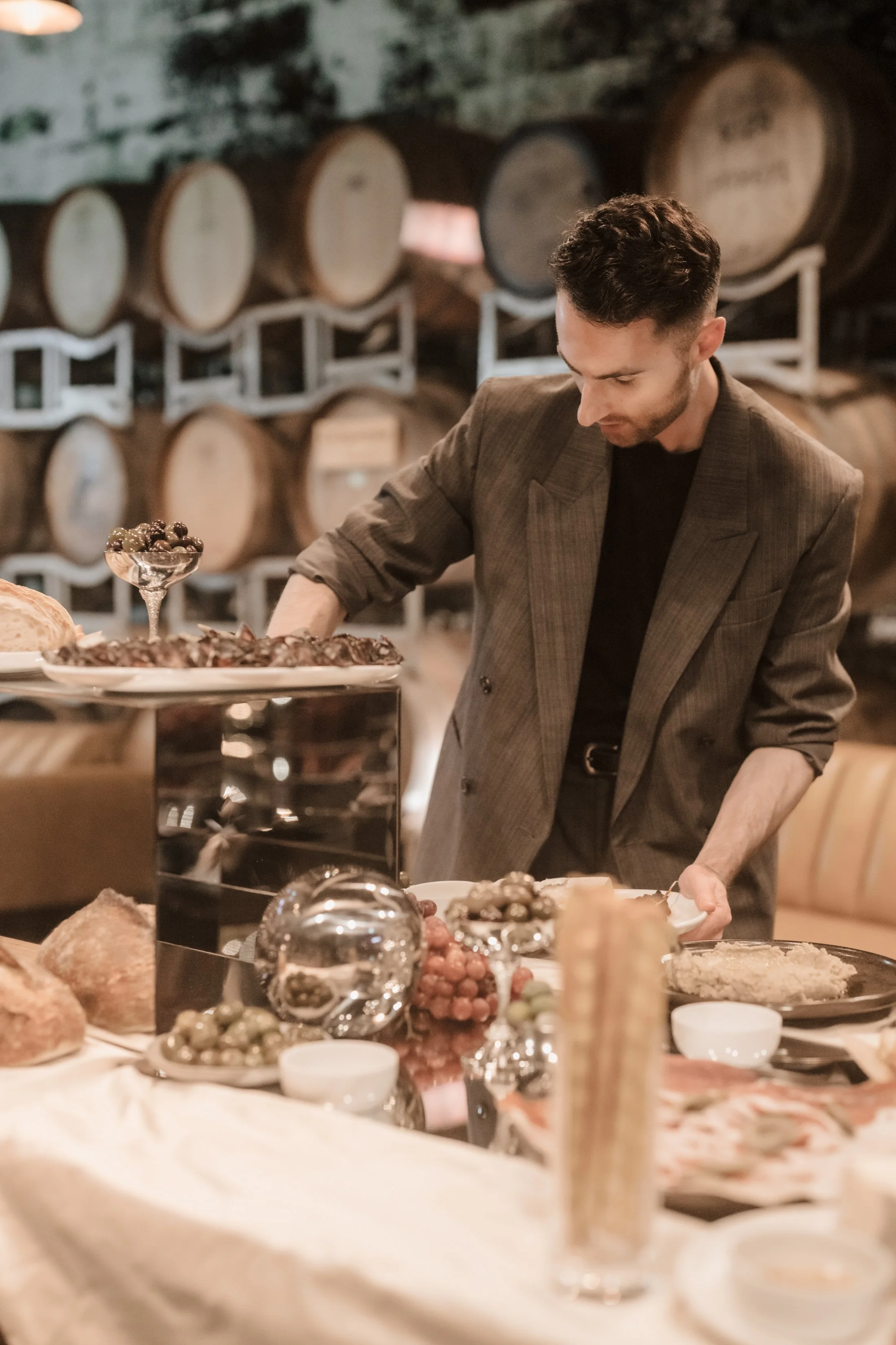 Alex Morabito, a stylish wedding planner, decorating a grazing table for a wedding with various foods, including grapes, bread, and cheese for Ever After Effect, in a rustic setting with wine barrels in the background at The Epicurean Red Hill.