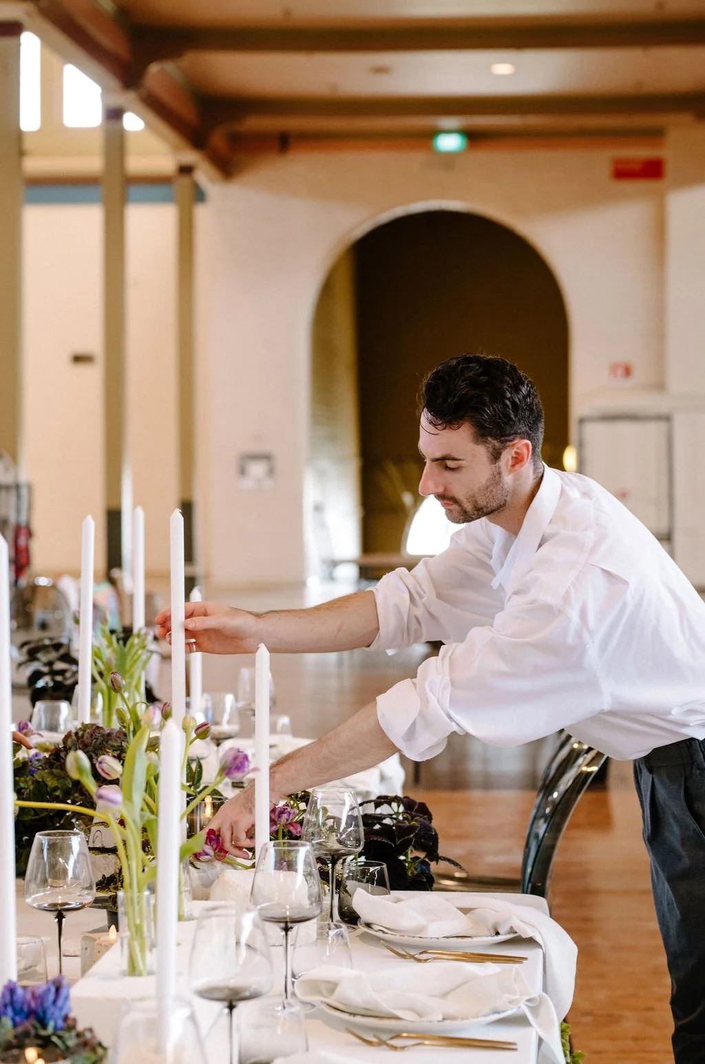 A man in a white shirt arranging a floral centerpiece on a table set for a formal dinner with wine glasses, plates, and napkins in a decorated event space.