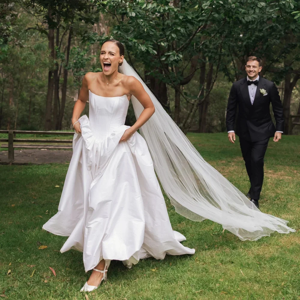 A bride in a white wedding dress laughing and walking outdoors, with a groom in a black tuxedo following her at The Cambium in Red Hill with green trees and grass after their wedding ceremony planned by Ever After Effect