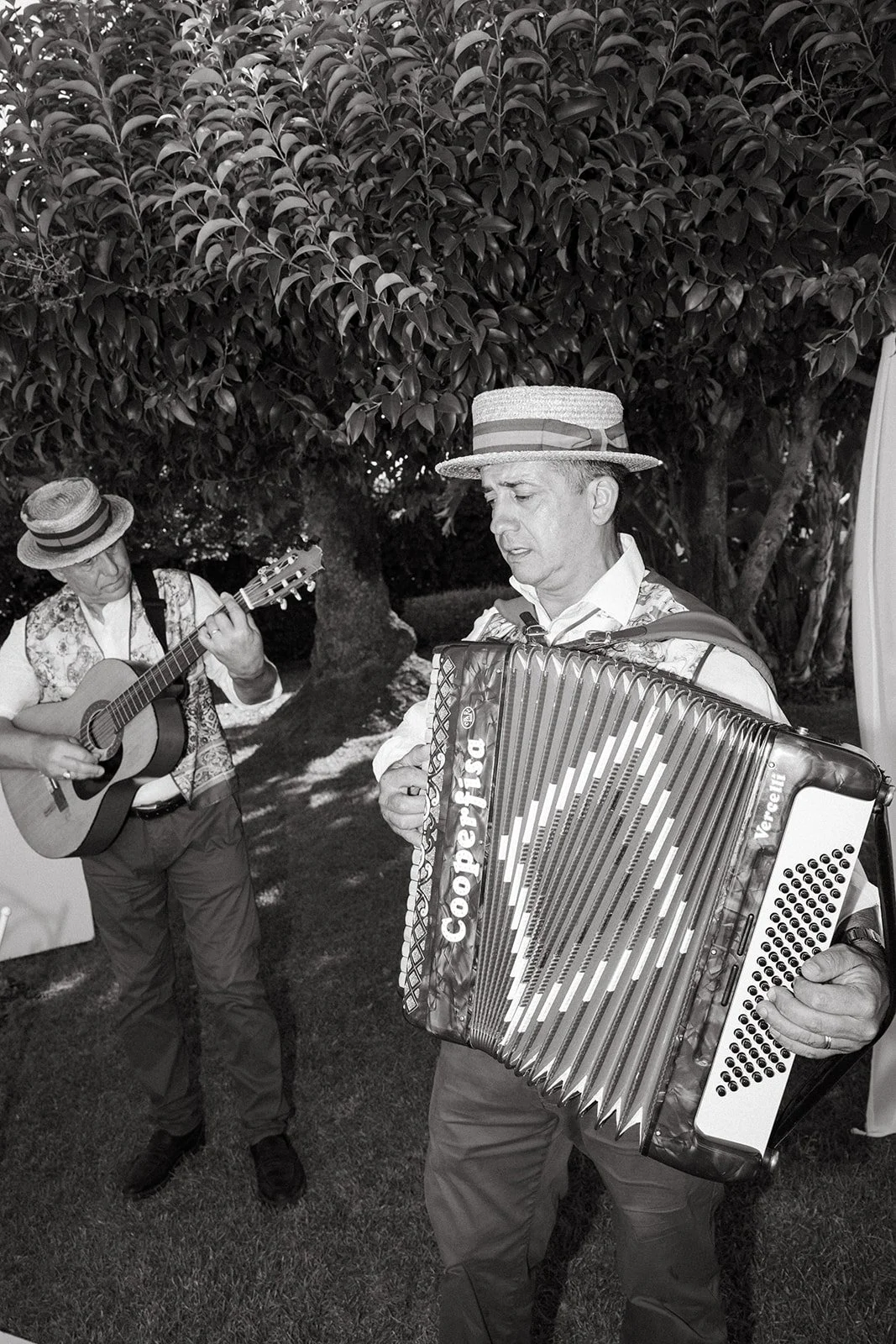 Two Italian musicians playing instruments outdoors near trees; one playing guitar, the other on an accordion, both wearing hats.