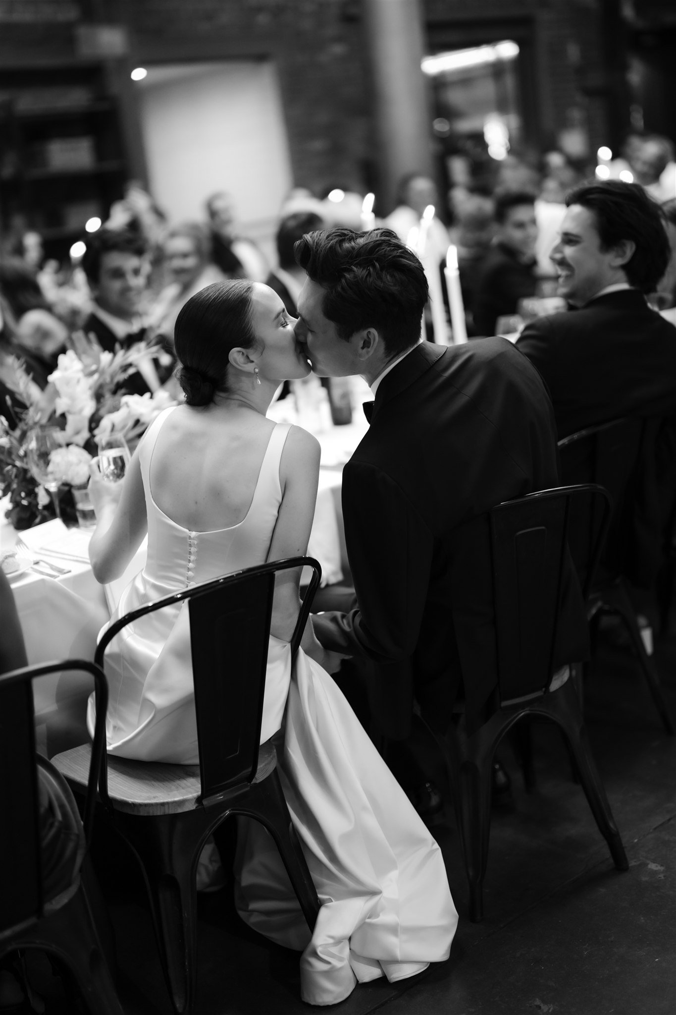 Black and white photograph of a bride and groom kissing at their wedding reception on the Mornington Peninsula. Guests are seated at a long table in the background, smiling and watching the couple.
