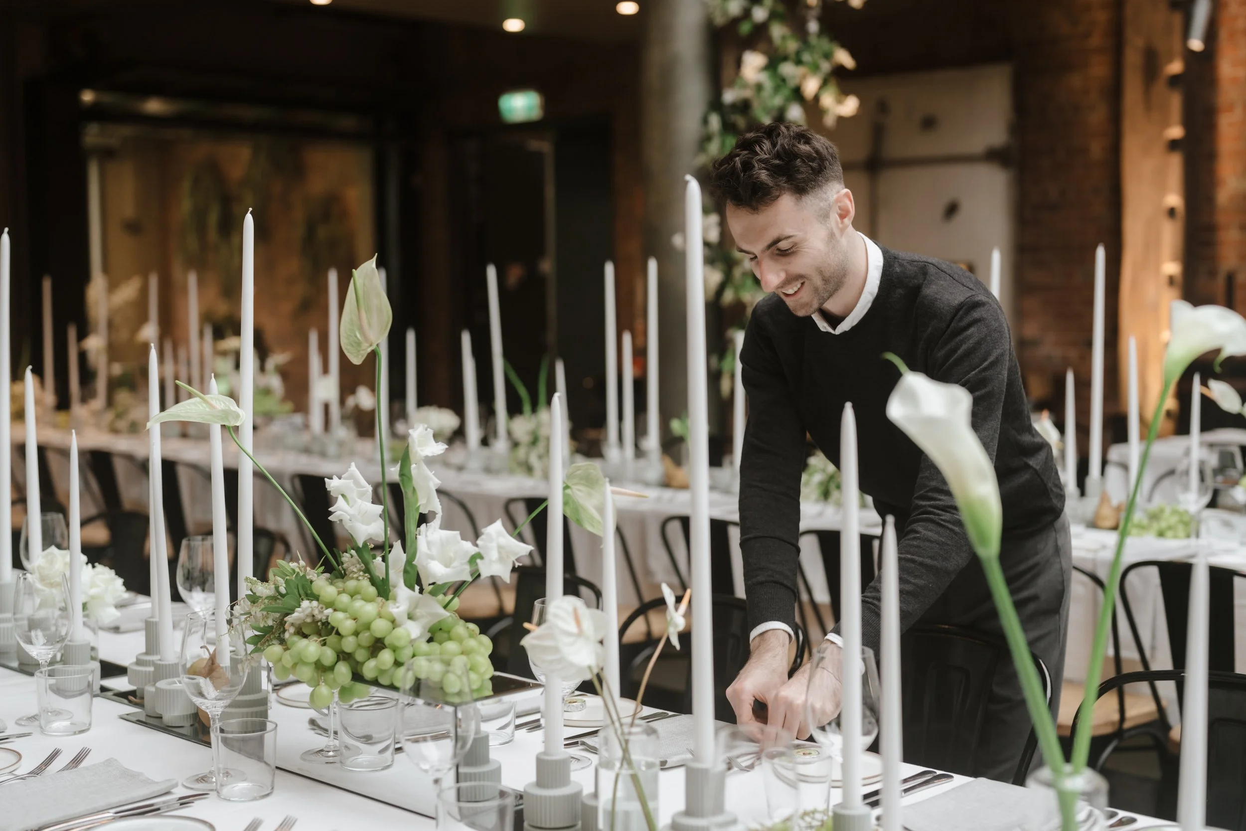 Alex Morabito arranging and styling a table decorated with white flowers, green grapes, and tall white candles at The Epicurean for Ever After Effect.
