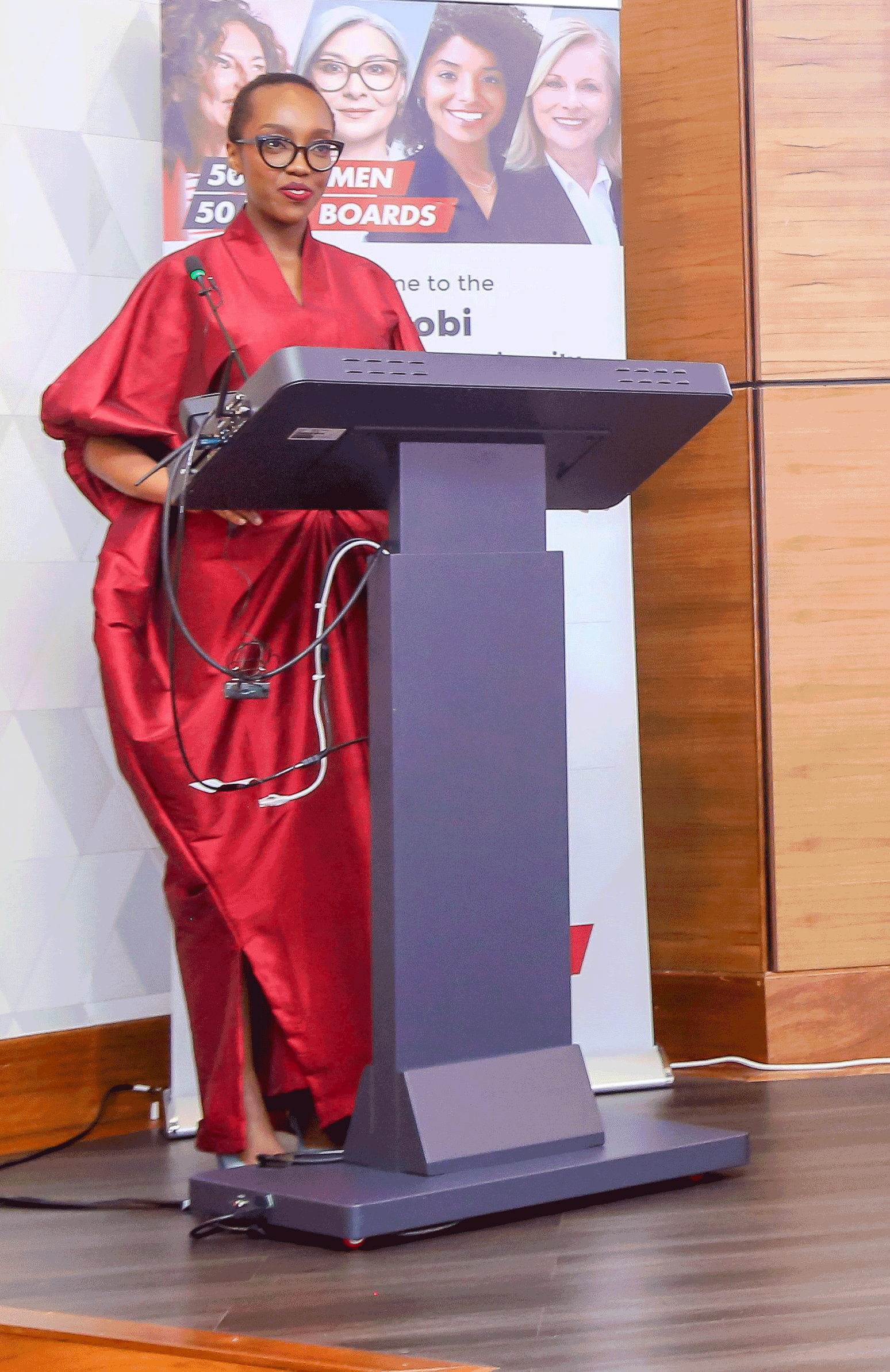 A woman wearing a red traditional dress and glasses is standing at a podium, speaking at an event. Behind her is a poster displaying images of five women and text related to women on boards.