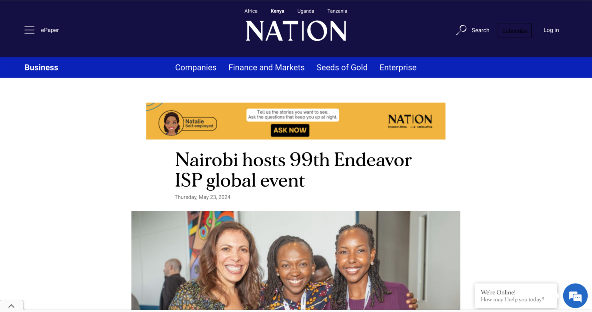 Photograph of three smiling women at a professional event, two of them are wearing patterned blouses and the third woman is wearing a dark blazer, standing indoors with a colorful abstract painting in the background.
