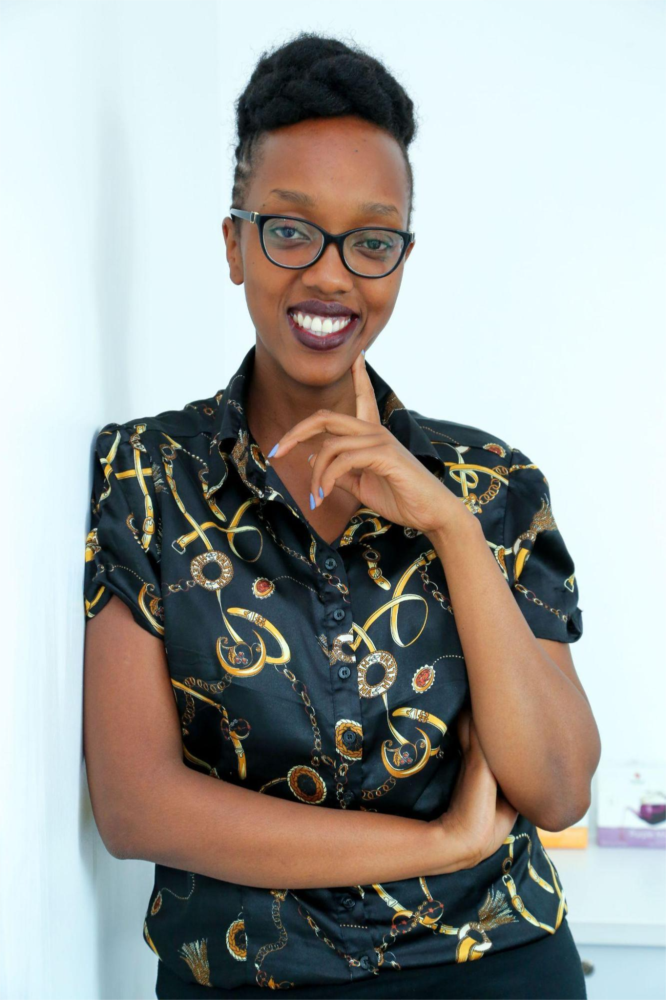 A smiling woman with glasses, wearing a patterned black shirt, standing indoors against a white background.