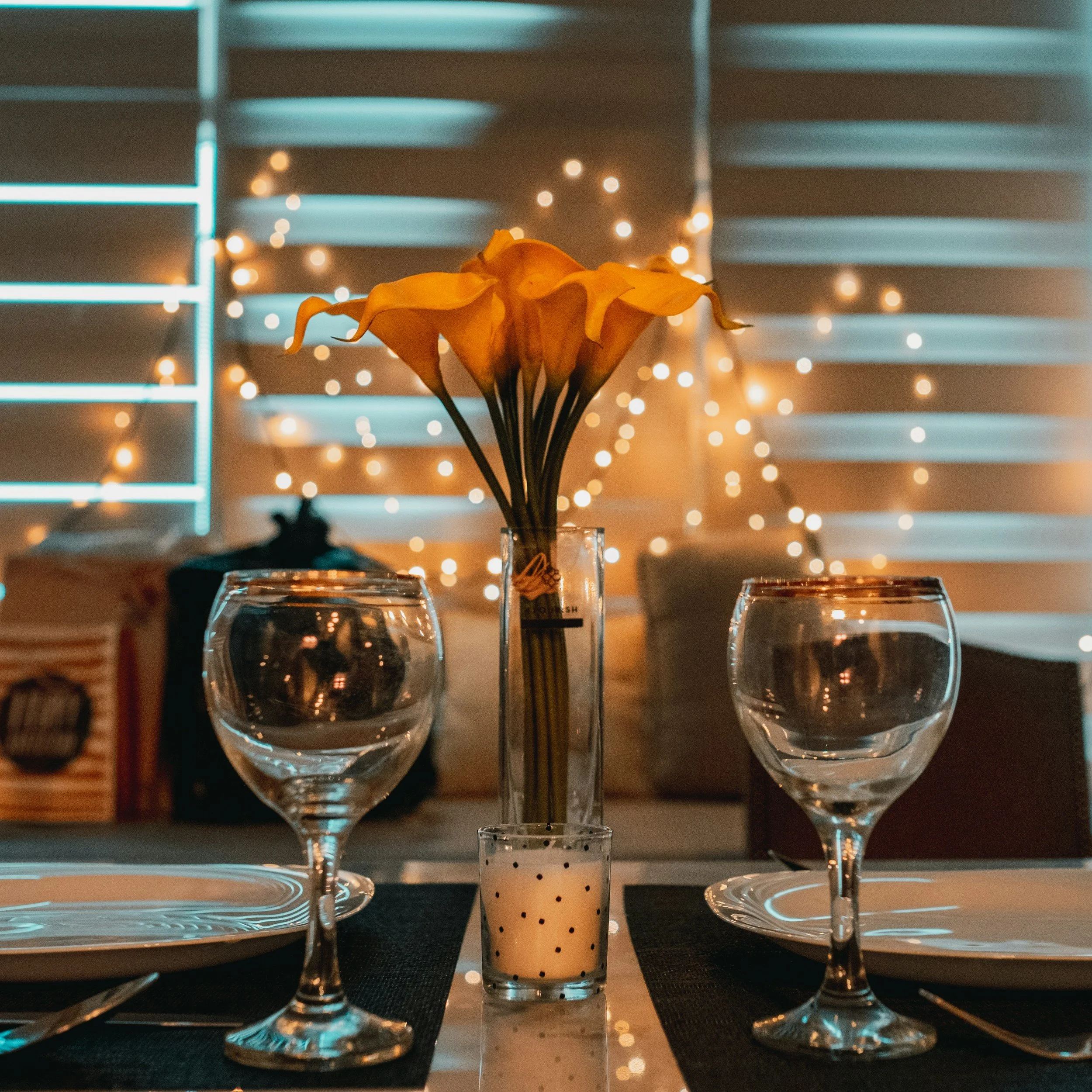 A dinner table set with two water glasses, a vase with yellow calla lilies, and a small candle, with string lights and shutters in the background.