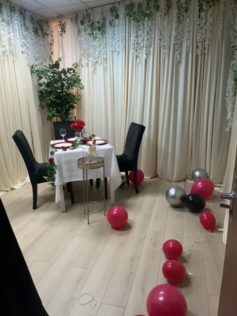 A decorated dining area for a romantic occasion with a small round table covered with a white cloth, set with wine glasses, flowers, and candles. Two black chairs are positioned around the table. The room has cream curtains, a potted plant, and scattered red, black, and silver balloons on the floor, with some curling ribbons.