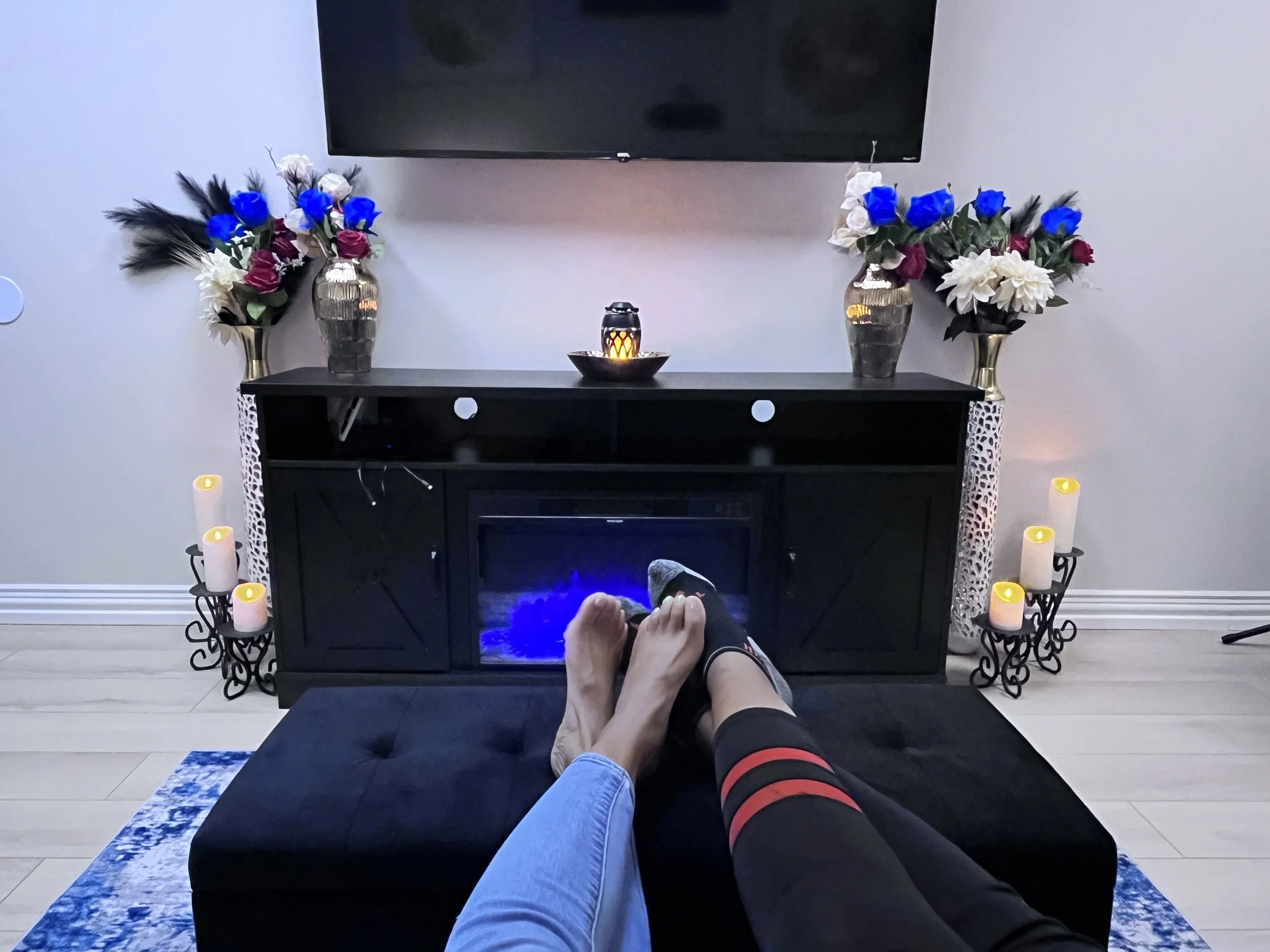 Two people lounging with their feet up on a black ottoman in front of a black entertainment console with a television above, decorated with two large vases of blue, white, and purple flowers, surrounded by lit candles on both sides.