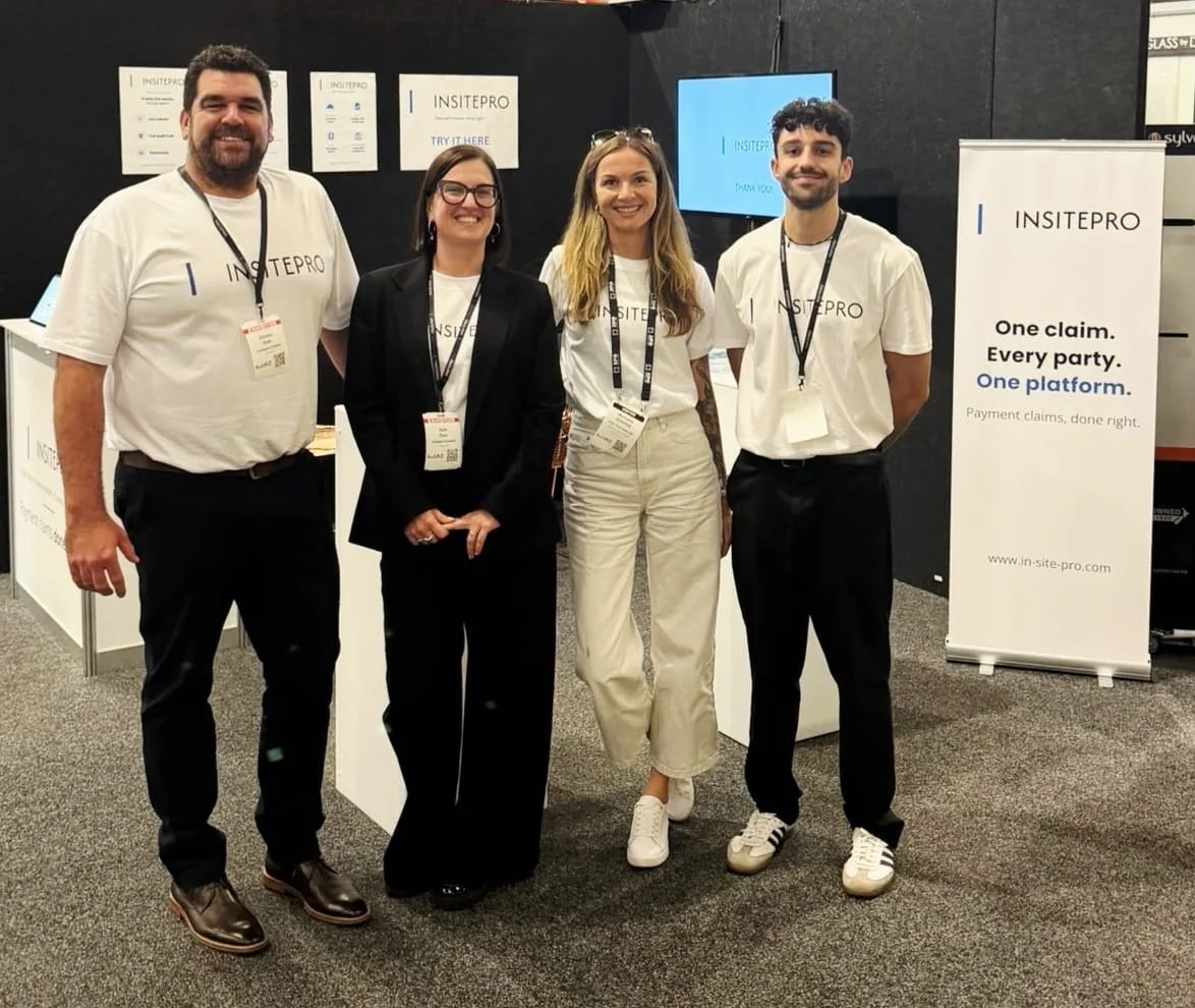 Four people standing together at an event booth for INSITEPRO, smiling at the camera. Two men and two women, three are wearing white T-shirts with the company's name, and one woman is wearing a black blazer. There are banners and screens behind them displaying the company's name and slogan.