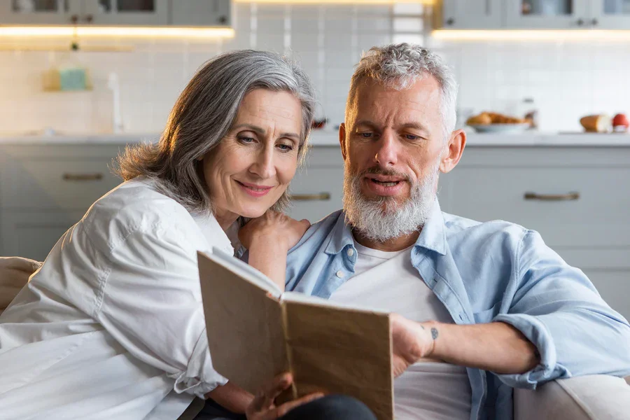 Older couple sitting on a sofa in a kitchen, reading a book together.