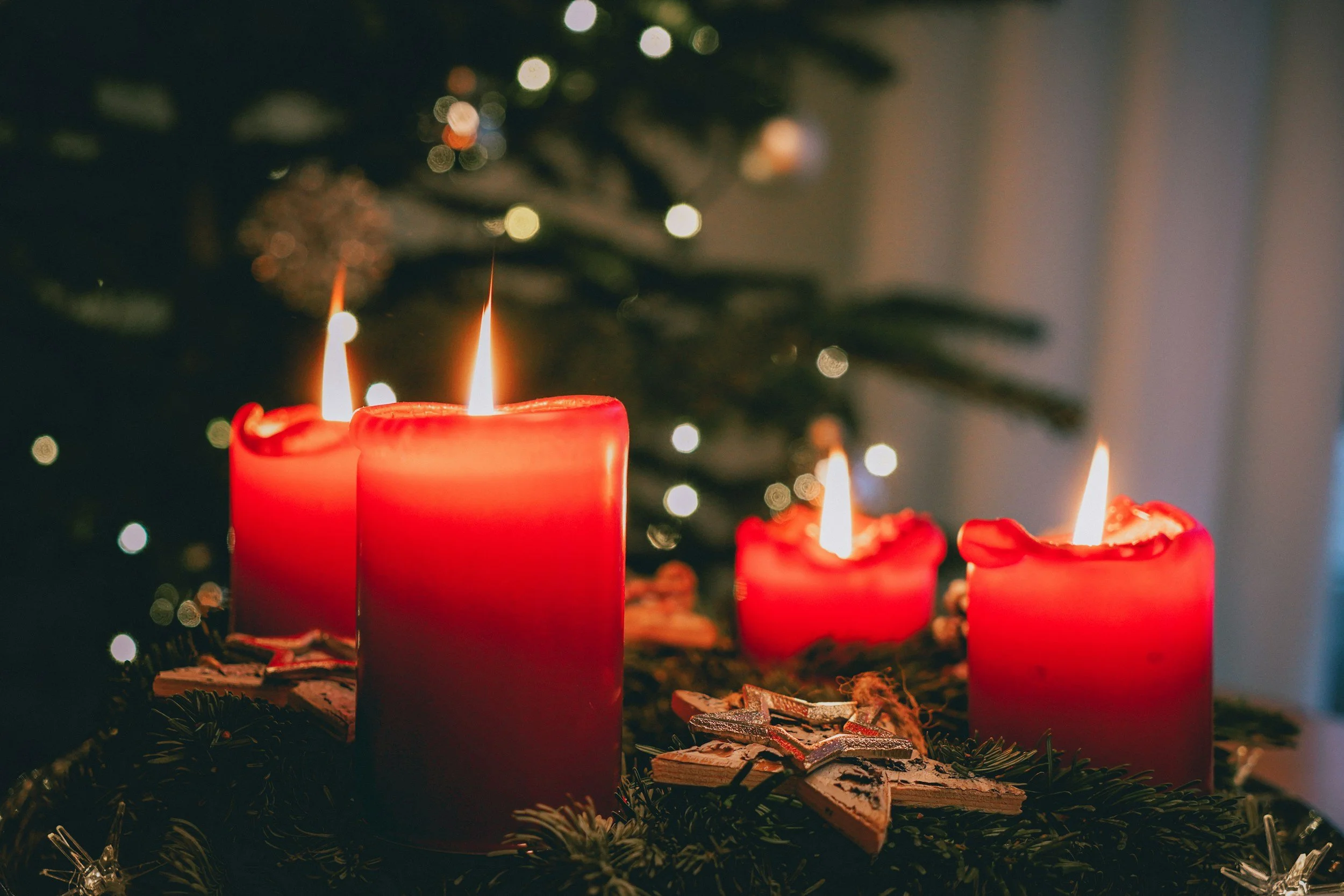 Three lit red candles on a decorated Christmas wreath with Christmas tree lights in the background.