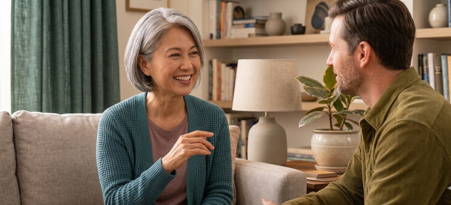 An older woman with short gray hair smiling and talking to a middle-aged man with brown hair and a beard in a cozy living room.