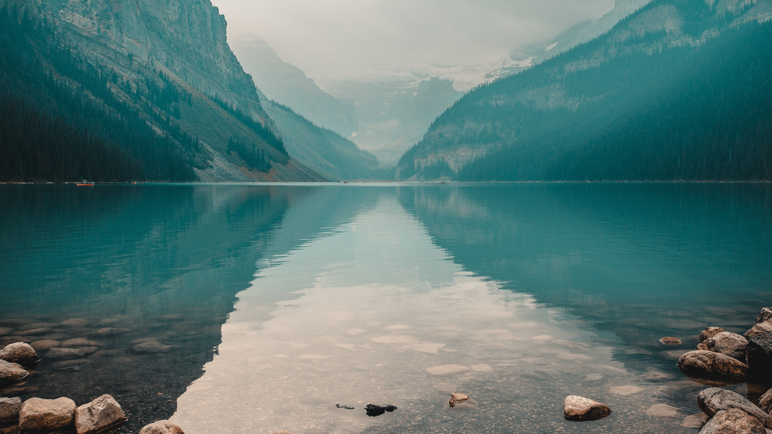 Calm mountain lake with mirror-like water, surrounded by lush green forests and steep cliffs, with a misty mountain backdrop