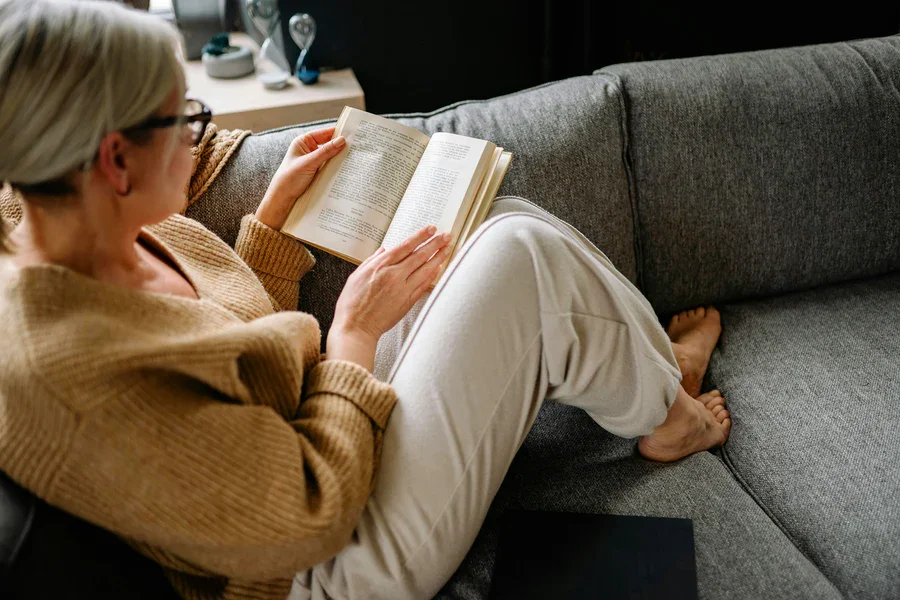 An older woman with glasses and gray hair reading a book while sitting on a couch with her legs crossed.