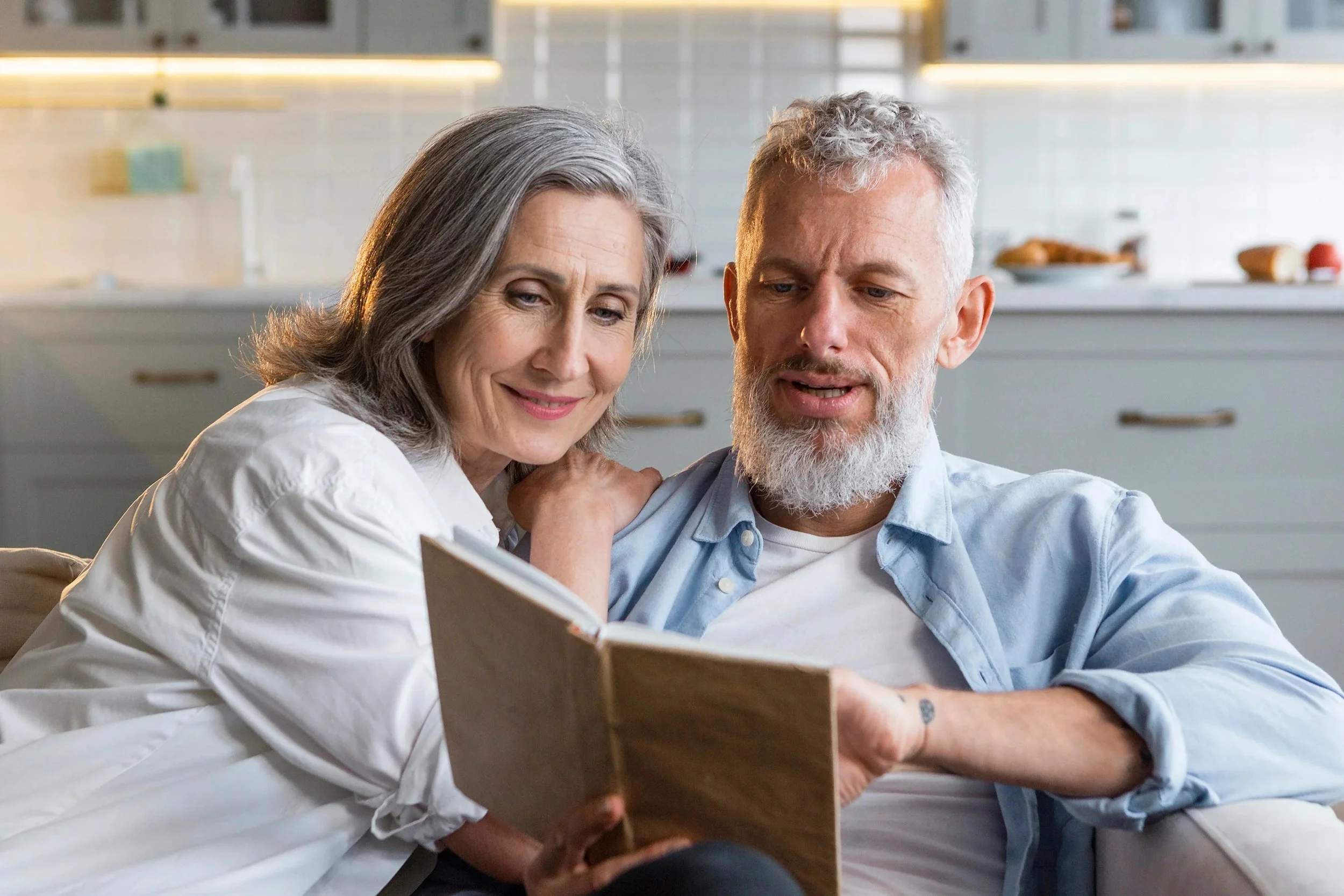 Older couple sitting close together indoors, reading a book or photo album, smiling and enjoying each other's company.