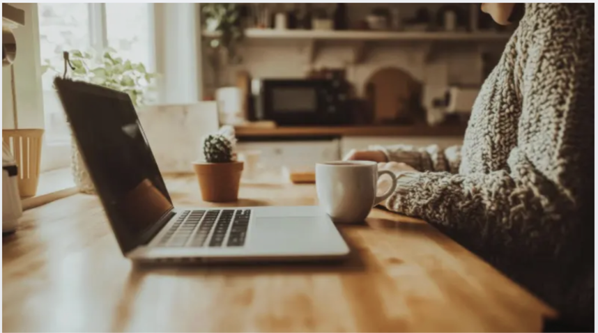 Person wearing a cozy sweater sitting at a wooden kitchen table, working on a laptop with a mug nearby, in a bright, homey kitchen.