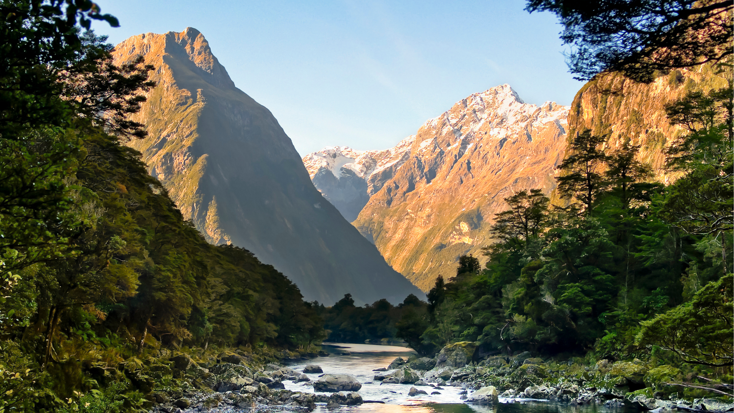 Mountain landscape with snow-capped peaks, dense forest, and a flowing river in the foreground.