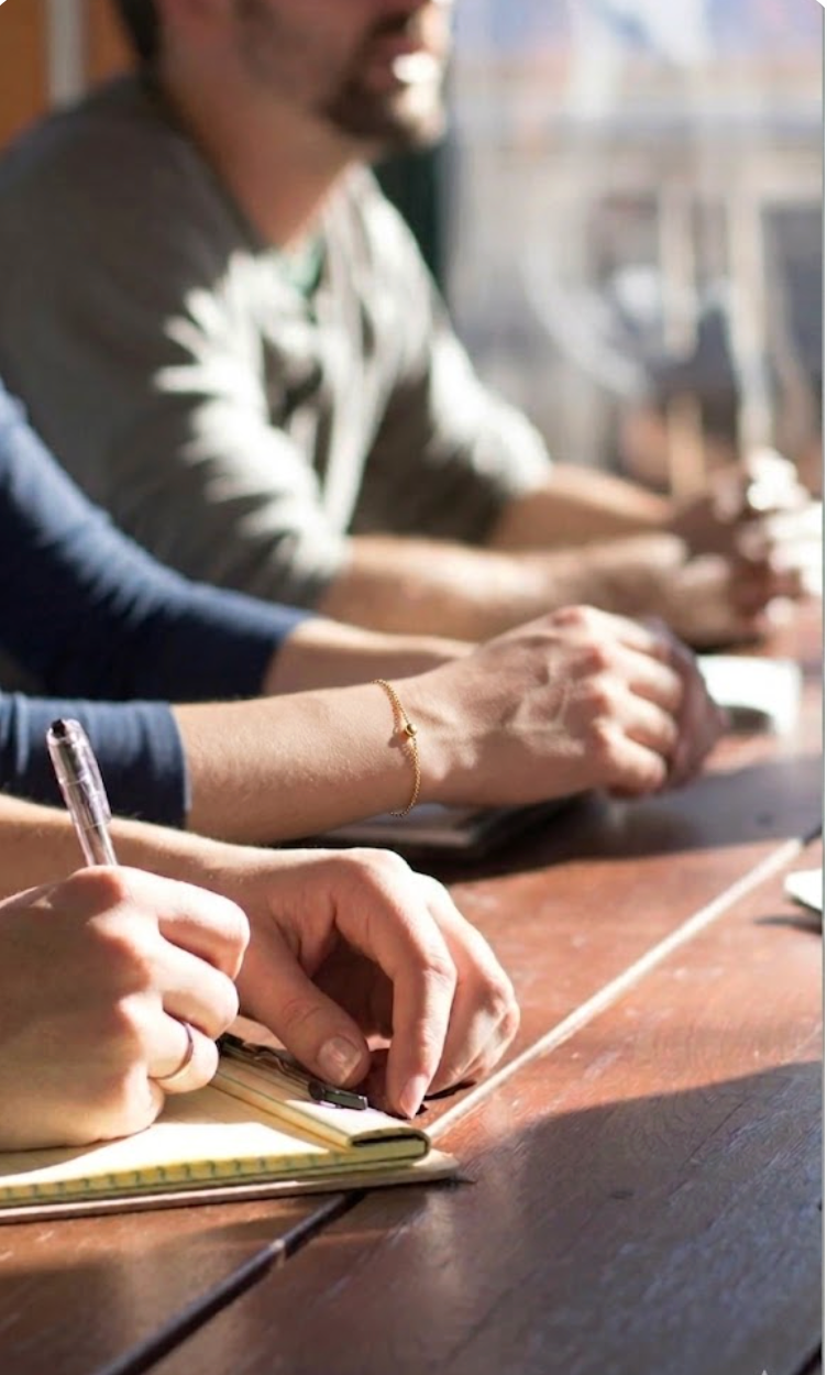 People seated at a table during a meeting, writing notes with notebooks and pens; sunlight streaming in.