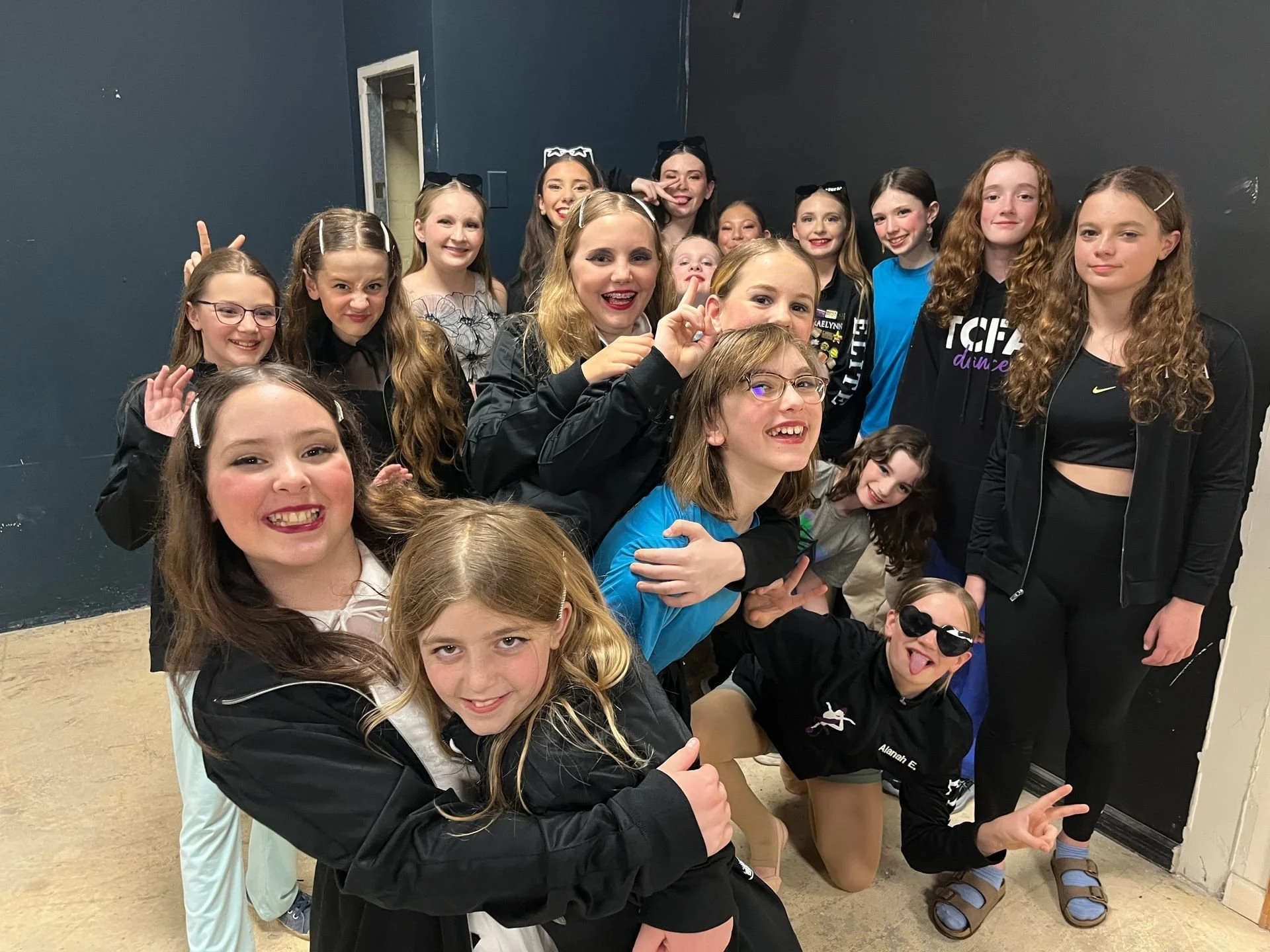 Group of smiling young girls and women posing together indoors, some making playful gestures, in front of a dark-colored wall.