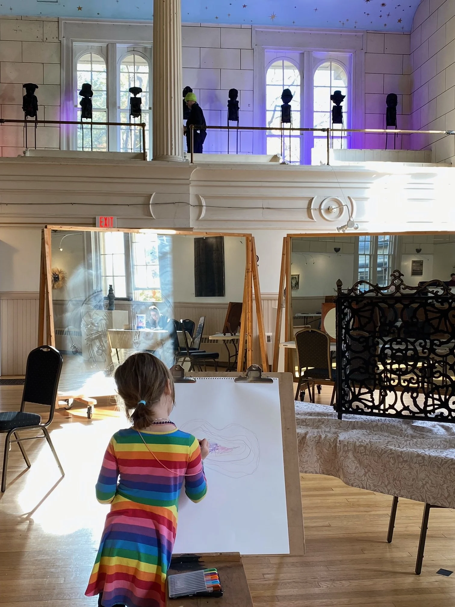A young girl with a rainbow-colored dress and ponytail is drawing on a whiteboard in a spacious room. The room has wooden floors, large windows, and a second-floor balcony with a decorative railing. Several mirrors and chairs are visible, along with a person wearing a green hat and a face mask, walking on the balcony. The ceiling is painted with stars and a blue sky.