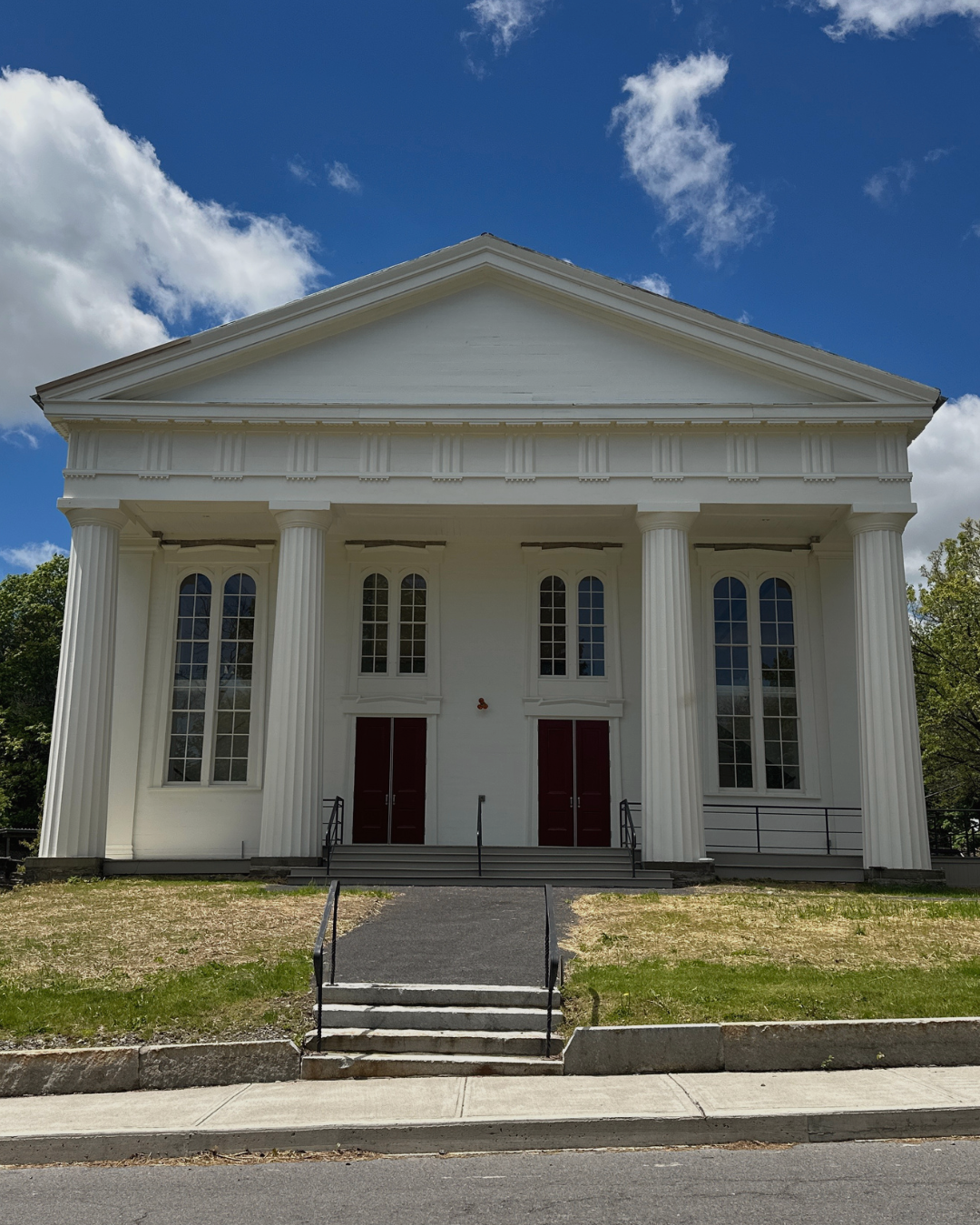 White neoclassical building with tall columns, arched windows, and red double doors, set against a bright blue sky with scattered clouds.