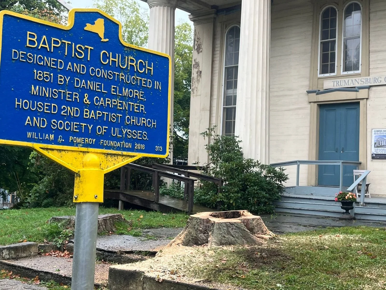 Blue historical marker in front of an old white church with columns and a blue door. The marker provides information about the church's history, built in 1851 by Daniel Elmore, and its connection to the Baptist church and society of Ulysses.