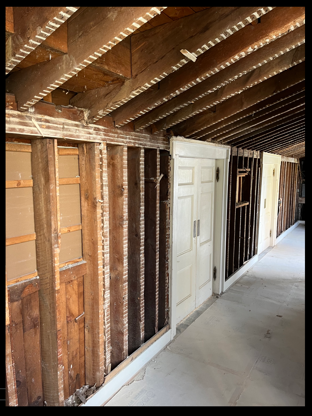 Interior view of renovation at TCFA with exposed wooden framing, a partially exposed lateral brick wall, and a white sliding door, along with an unfinished concrete floor.