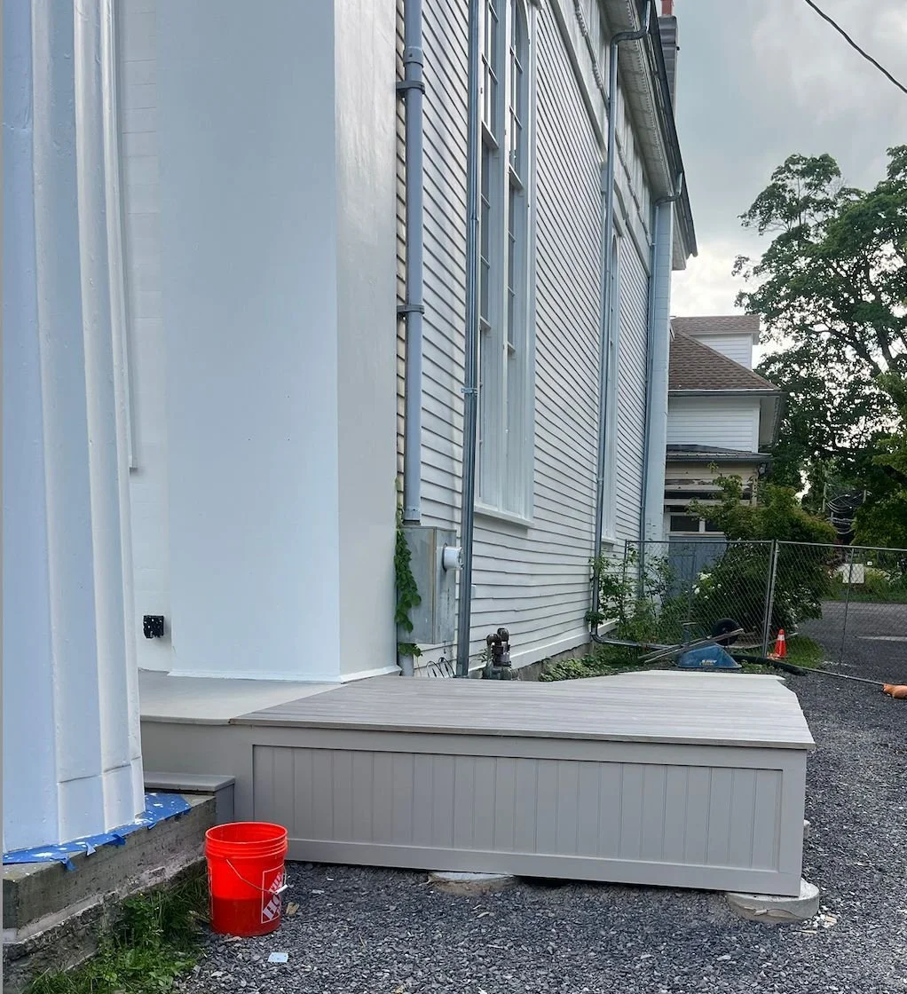 Backyard scene showing TCFA with vinyl siding and windows, a grey storage bench on a gravel surface, a red bucket, various pipes and wires, a chain-link fence, and trees in the background.