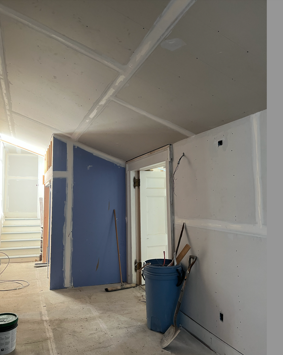 Interior of a the entry way under construction with unfinished drywall, a blue wall, and construction tools including a shovel and a bucket.