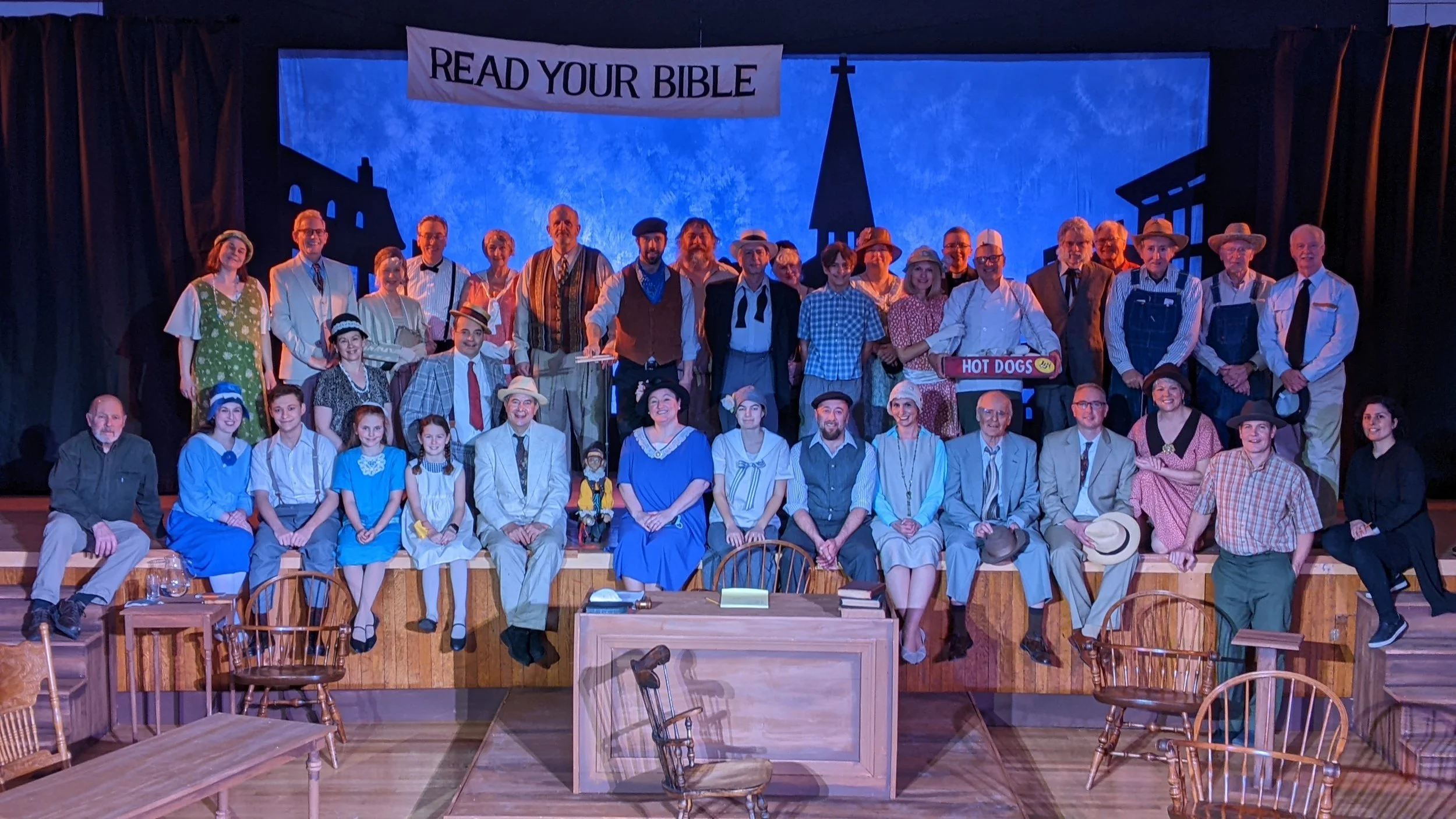Group of people on stage dressed in vintage clothing with a sign that says 'Read Your Bible,' historical and theatrical costumes, some sitting and some standing, in a theater setting.