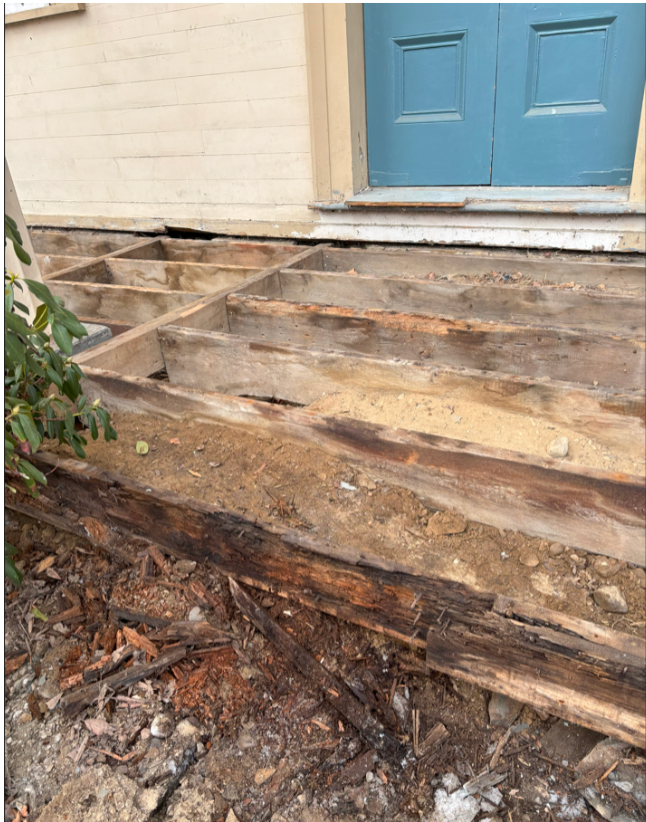 The image shows a damaged wooden staircase in front of a house, with some steps missing or broken, exposing dirt and debris underneath. To the right, there is a blue door and part of a white wall, with a green shrub on the left side.