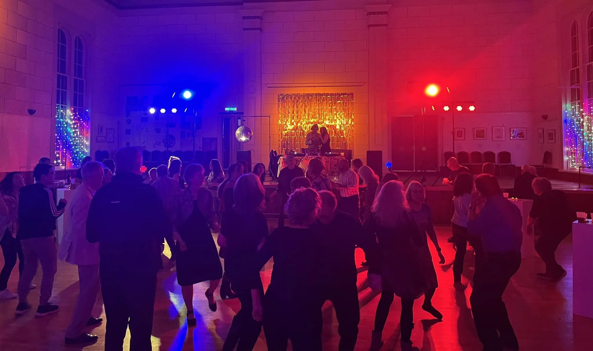 People dancing and socializing at a party with colorful lighting, a DJ booth, and a disco ball in a spacious hall.
