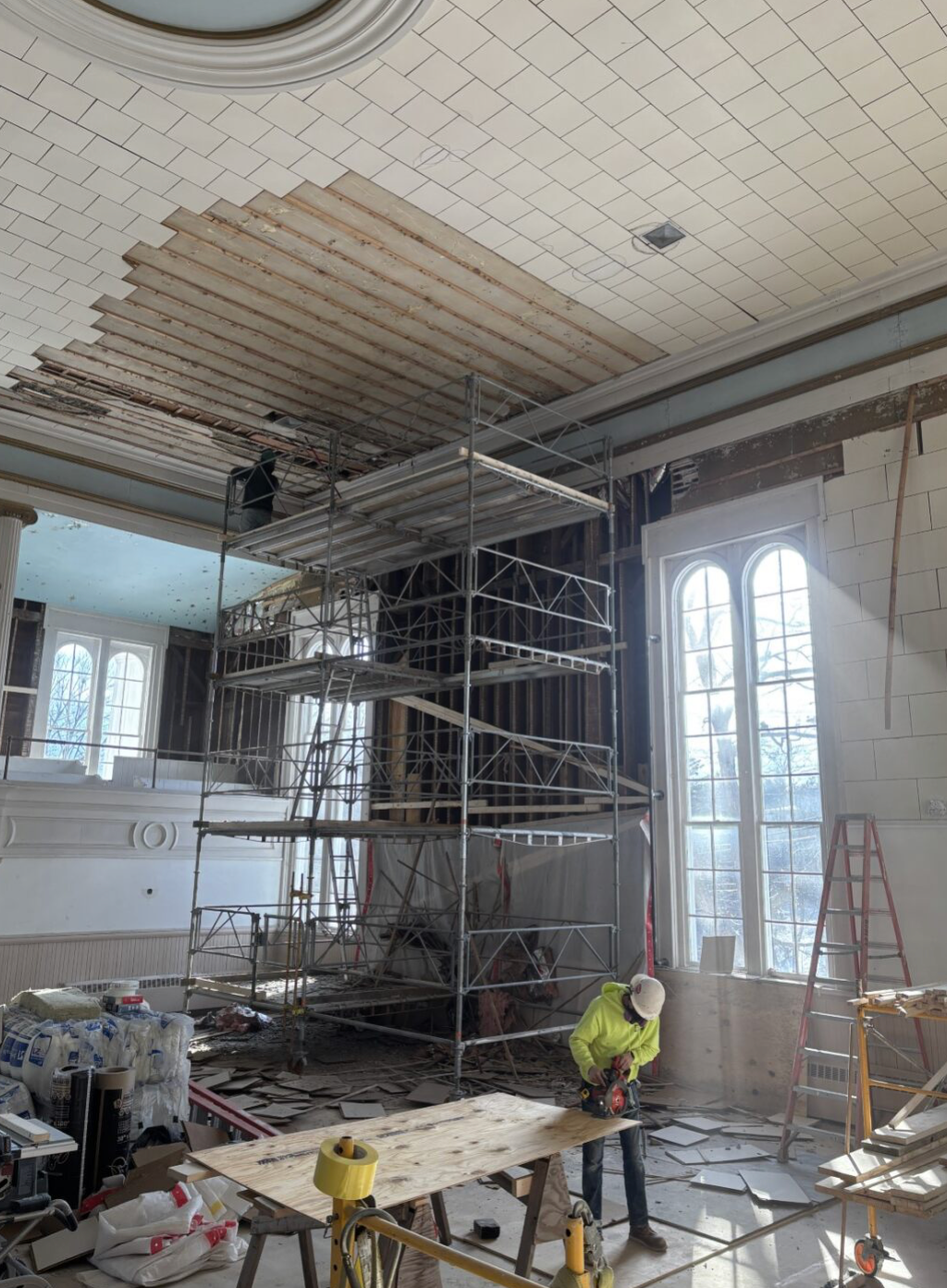 Construction workers renovating a large room with scaffolding, where the ceiling tiles are being replaced or repaired.