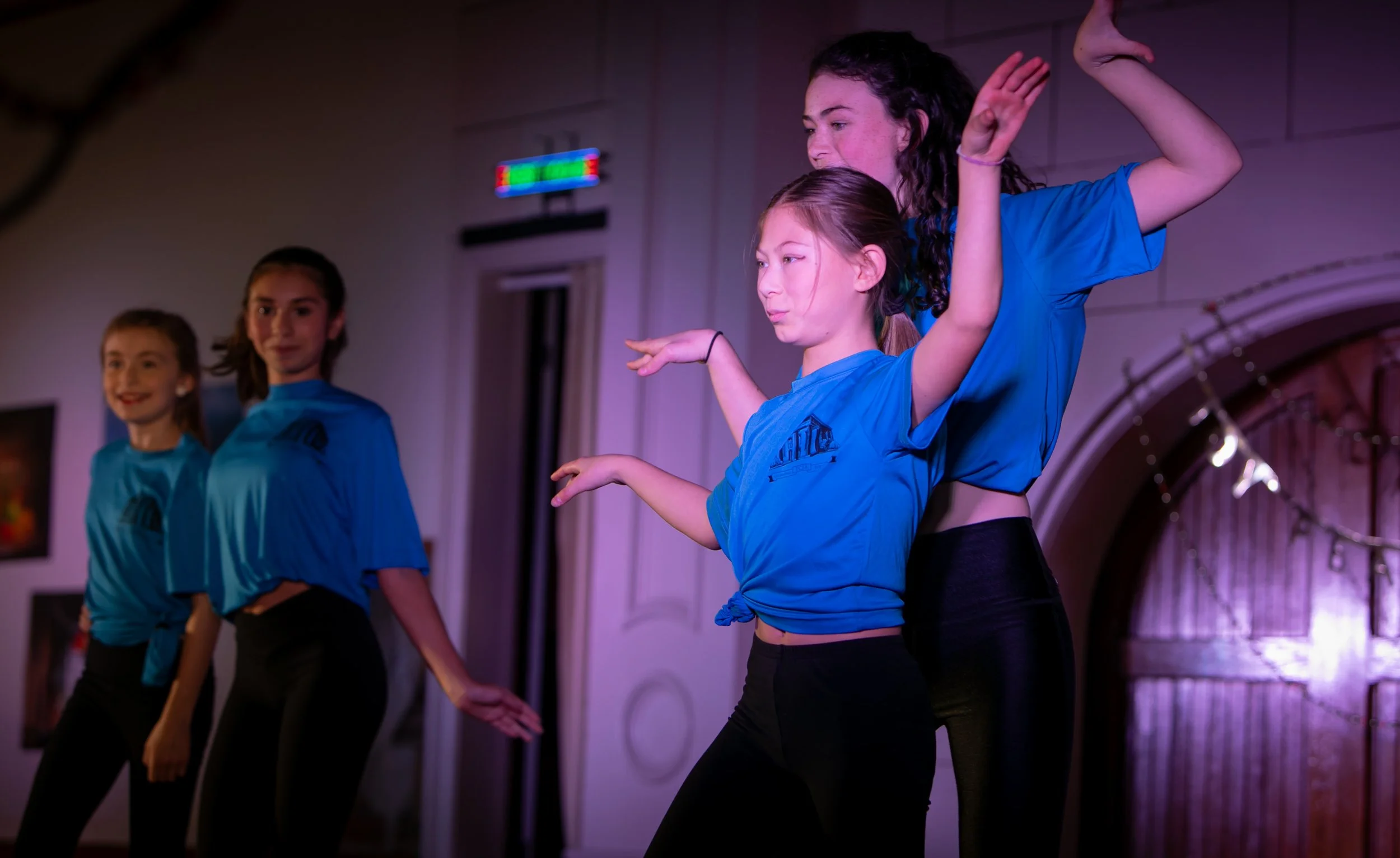 Four young girls dancing or performing on stage, wearing blue shirts and black pants, with a woman in black behind them, in a room with purple and pink lighting.