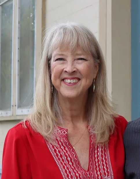 A woman with blonde hair smiling, wearing a red embroidered top, standing outdoors near a beige building and window.