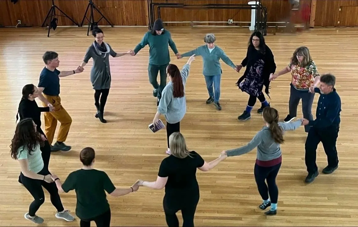 A group of people holding hands and dancing in a circle in a wooden-floored room.