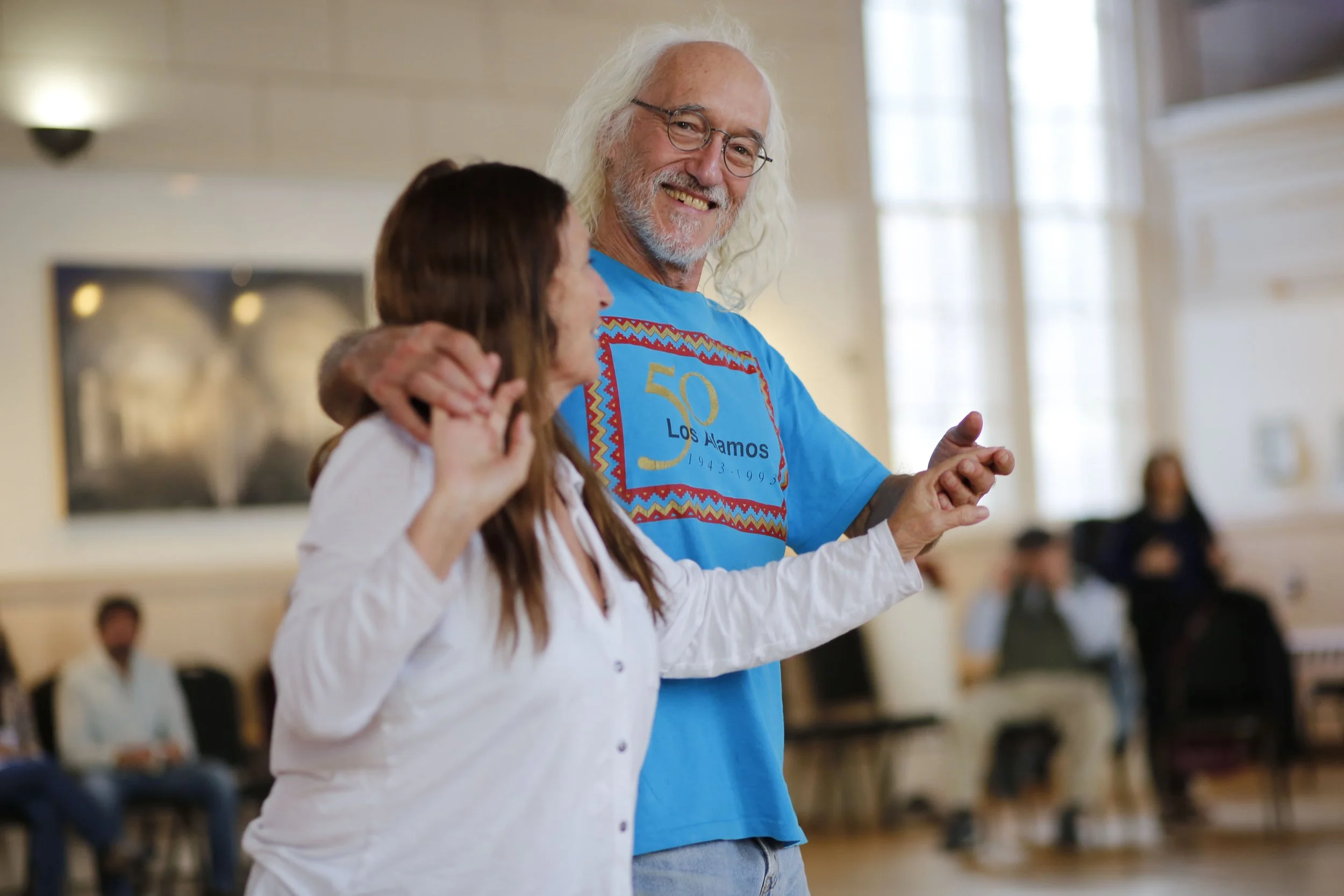 A smiling older man with long white hair and glasses wearing a blue T-shirt in a community gathering room, with a woman in a white shirt in the foreground. Several people are in the background.