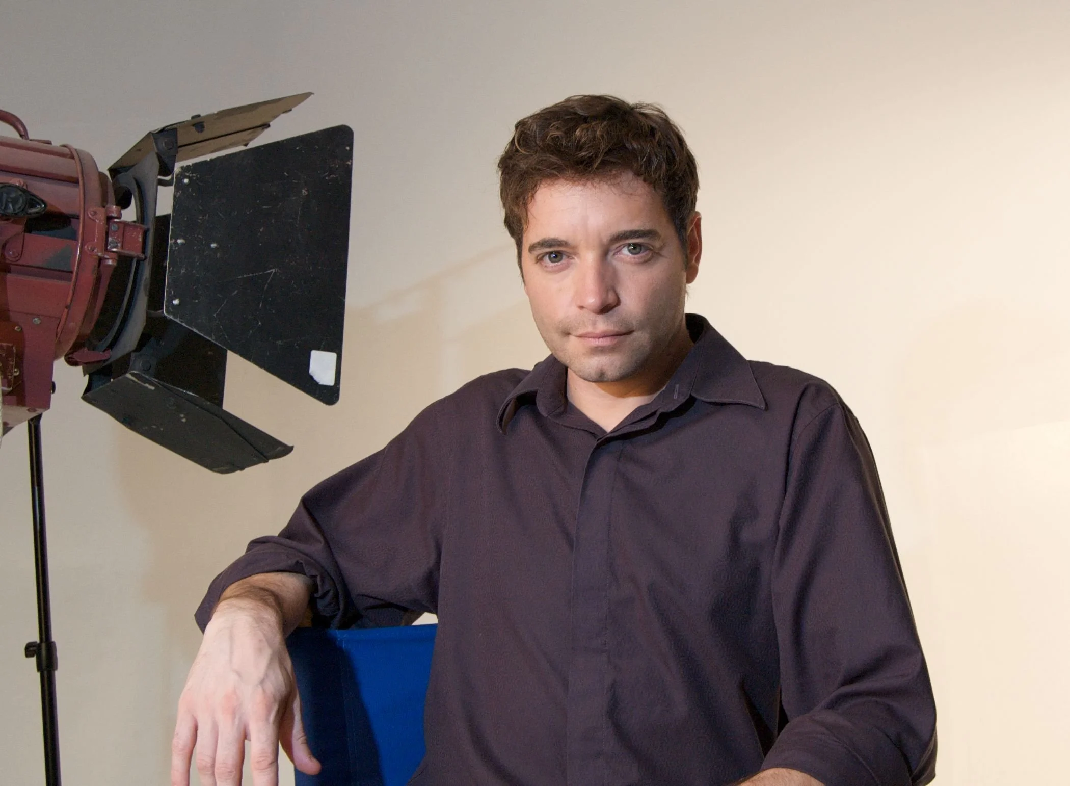 Picture of Danny Trippett with brown hair and light skin wearing a dark shirt, sitting in front of a light-colored wall, with film equipment nearby.