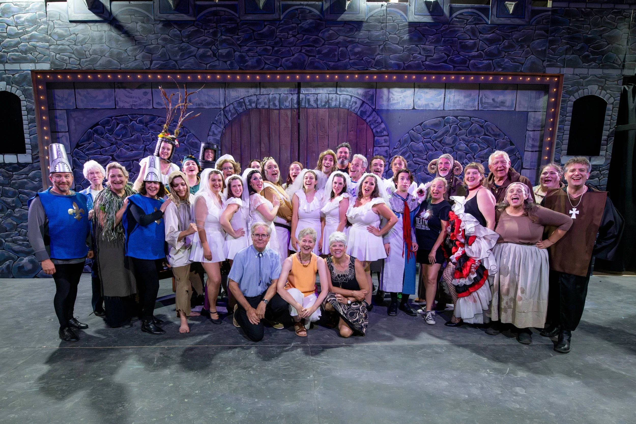 Group of theatrical performers and crew posing together on stage in costume after a play. Some are smiling and celebrating, with costumes ranging from medieval knights to fairy tale characters. The stage backdrop depicts a castle wall with a large wooden door.