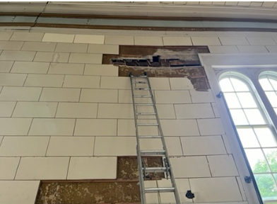 Interior wall with a large hole or damage, a metal ladder leaning against it, and a window with blinds to the right.