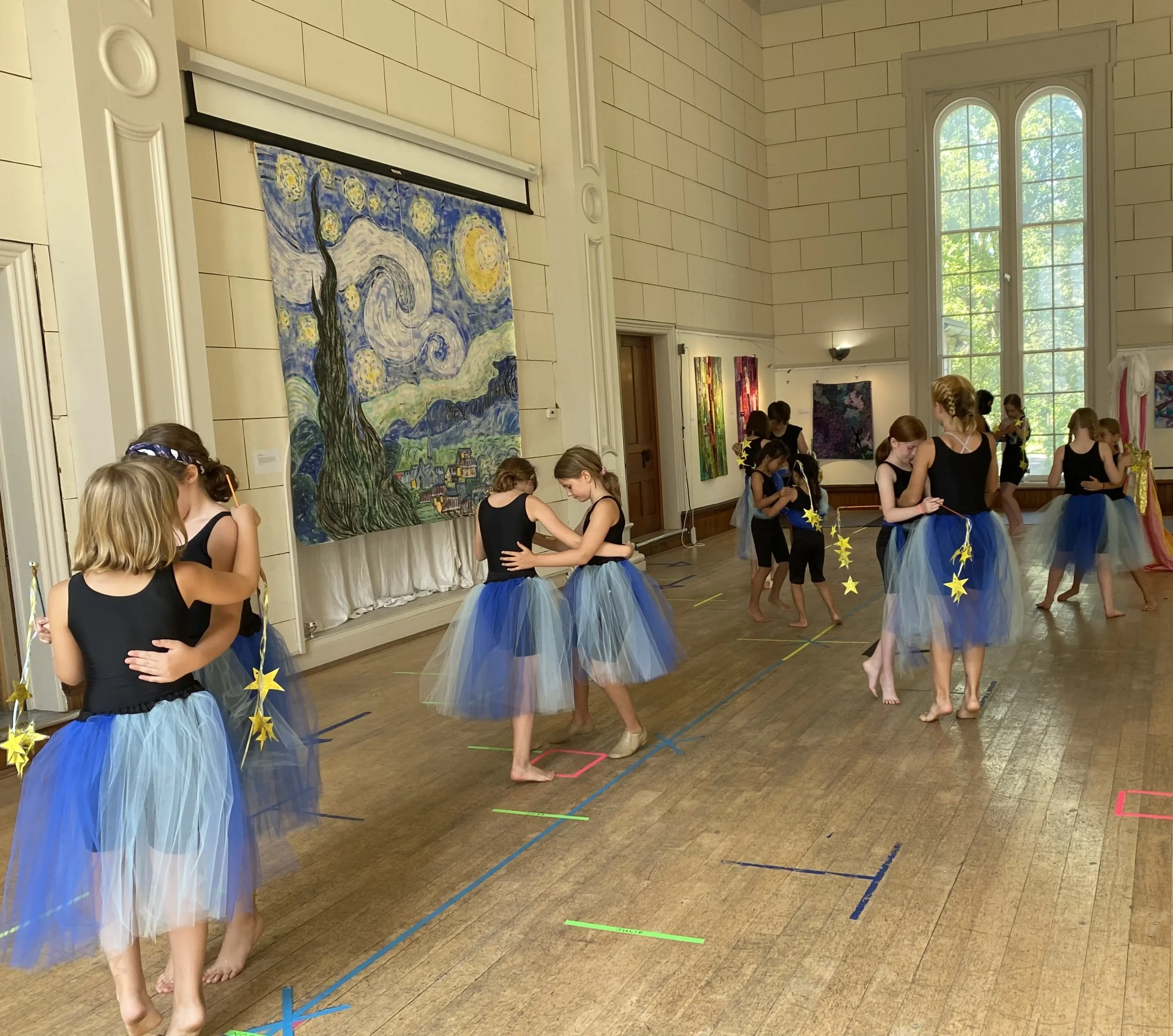 Children in ballet costumes practicing in a dance studio with large windows, artwork on the wall, and colorful tape on the floor.